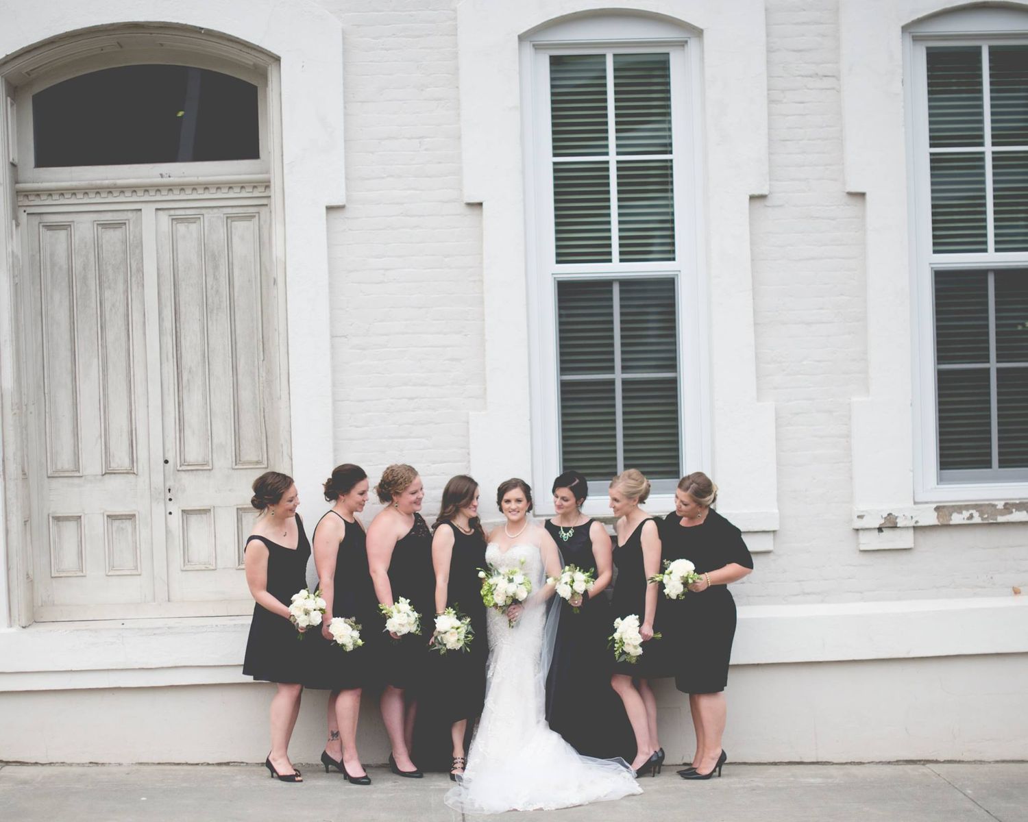 Bride surrounded by bridesmaids in black dresses holding white bouquets outside a white historic building.