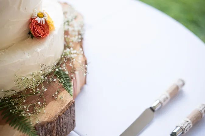 Rustic wedding cake on wood slice with wildflowers, ferns, and baby's breath, with a cake knife on white table.