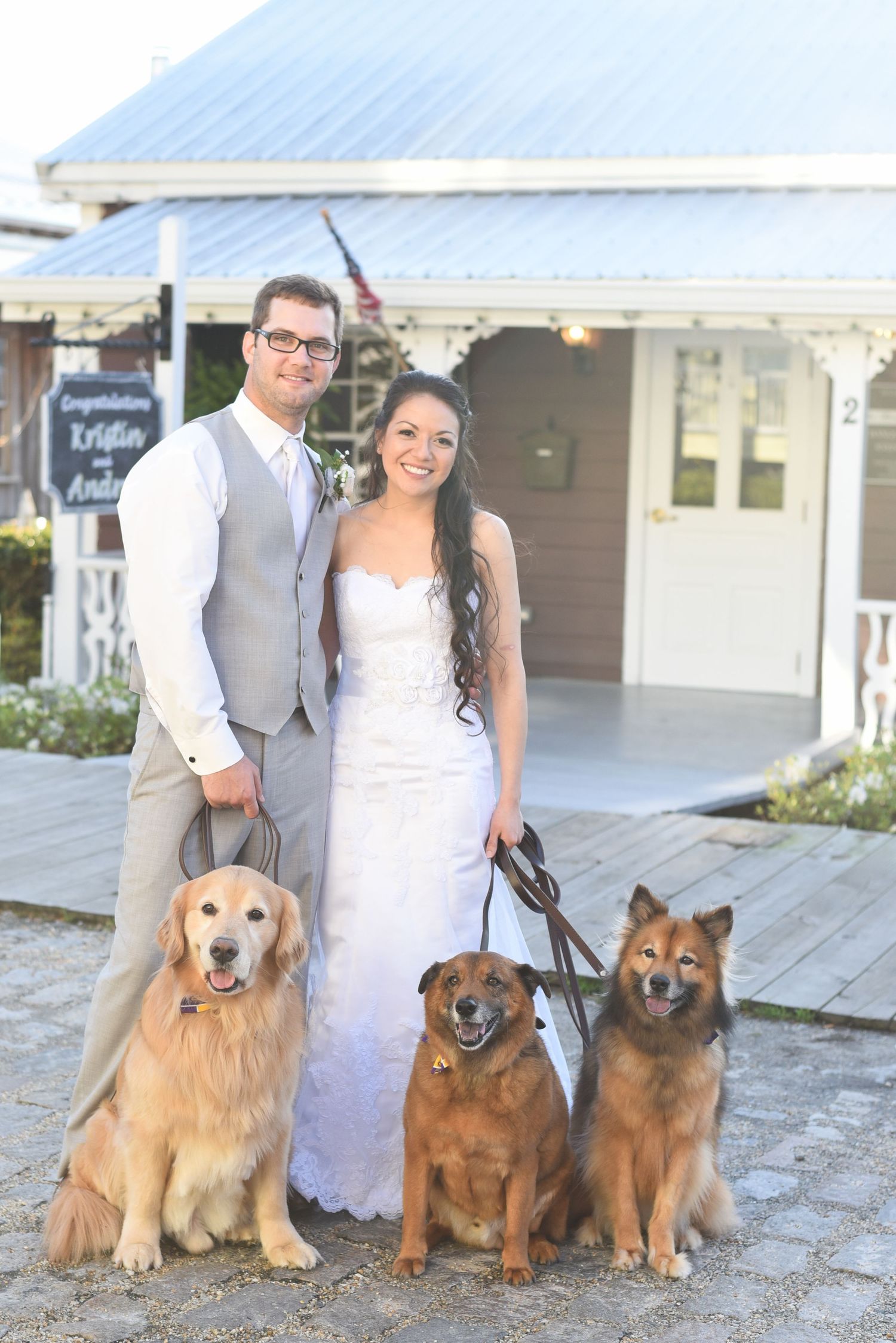 Smiling bride and groom pose outdoors with three dogs in front of a decorated wedding venue sign.