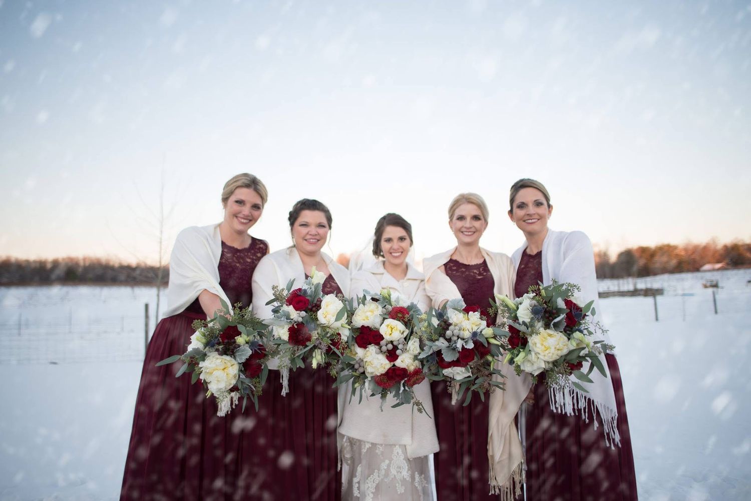 A bride and four bridesmaids in burgundy gowns pose with bouquets during a snowy winter wedding outdoors.