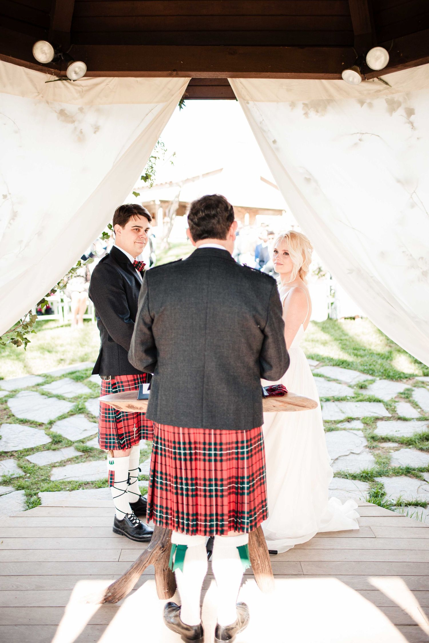 Outdoor wedding ceremony with groom in red tartan kilt and bride in white dress under a draped gazebo.
