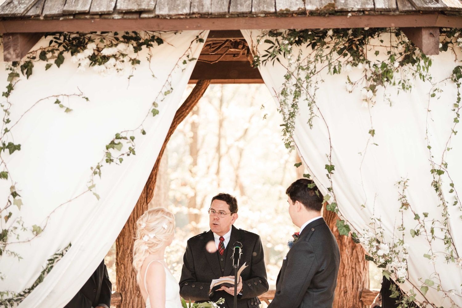 Two grooms exchange vows under a rustic wooden arch adorned with white draping and greenery at an outdoor wedding ceremony.