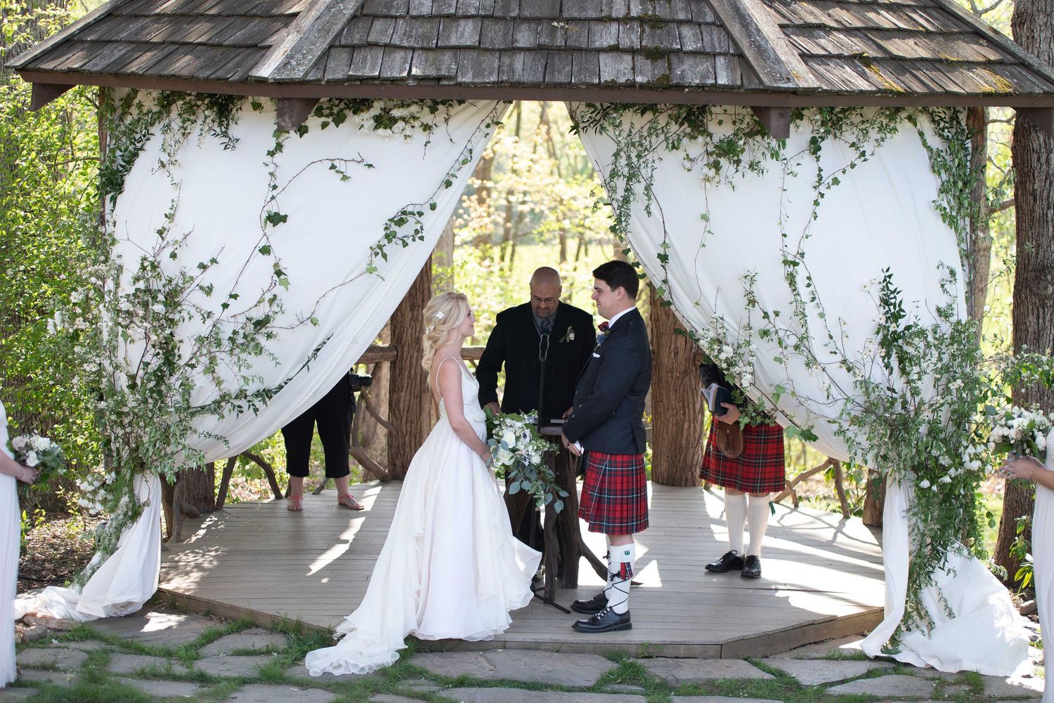 Bride and groom exchange vows under a floral ivy-draped gazebo at an outdoor wedding ceremony.