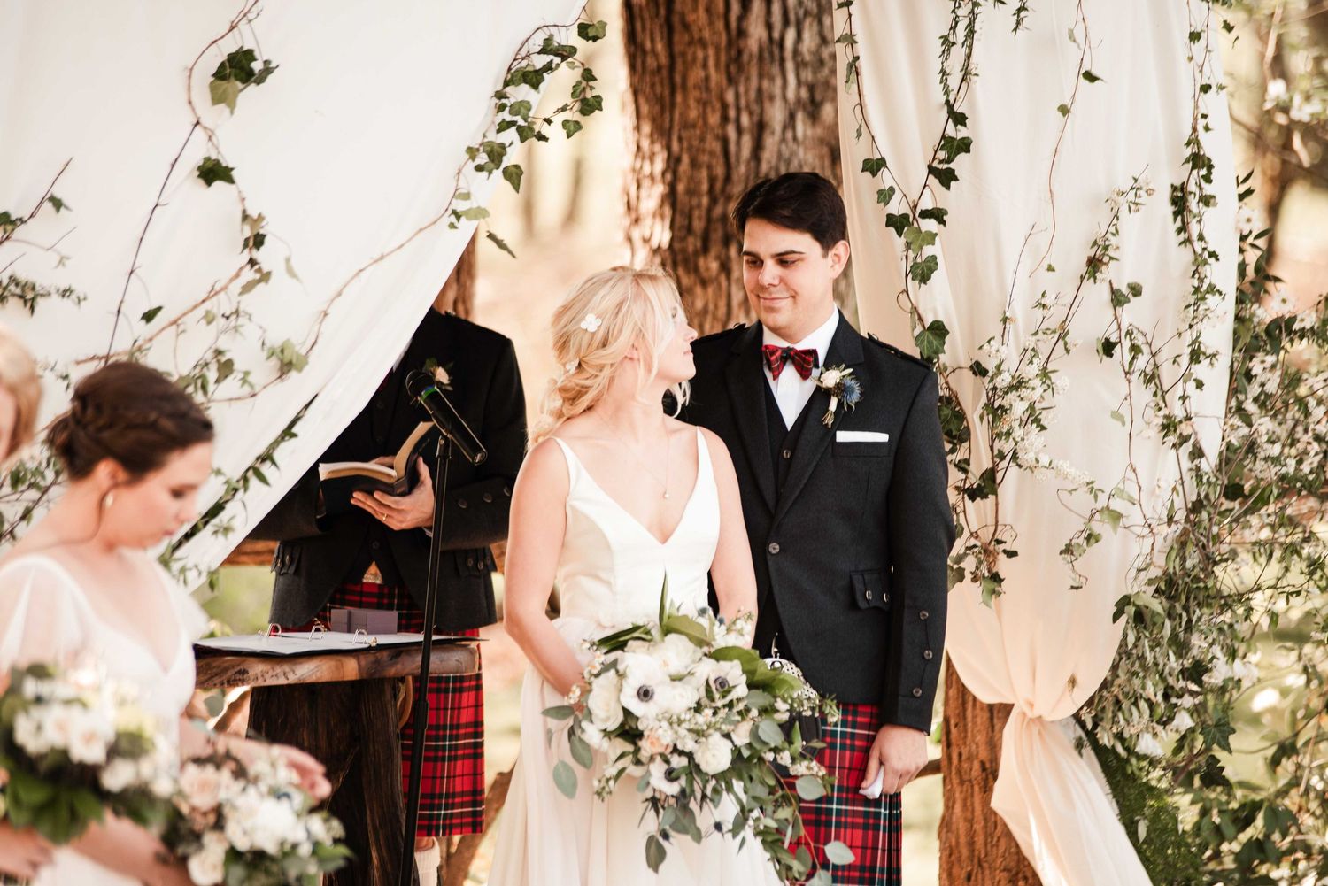 Bride and groom exchange vows at elegant wedding ceremony with greenery backdrop, groom wearing traditional Scottish tartan kilt.