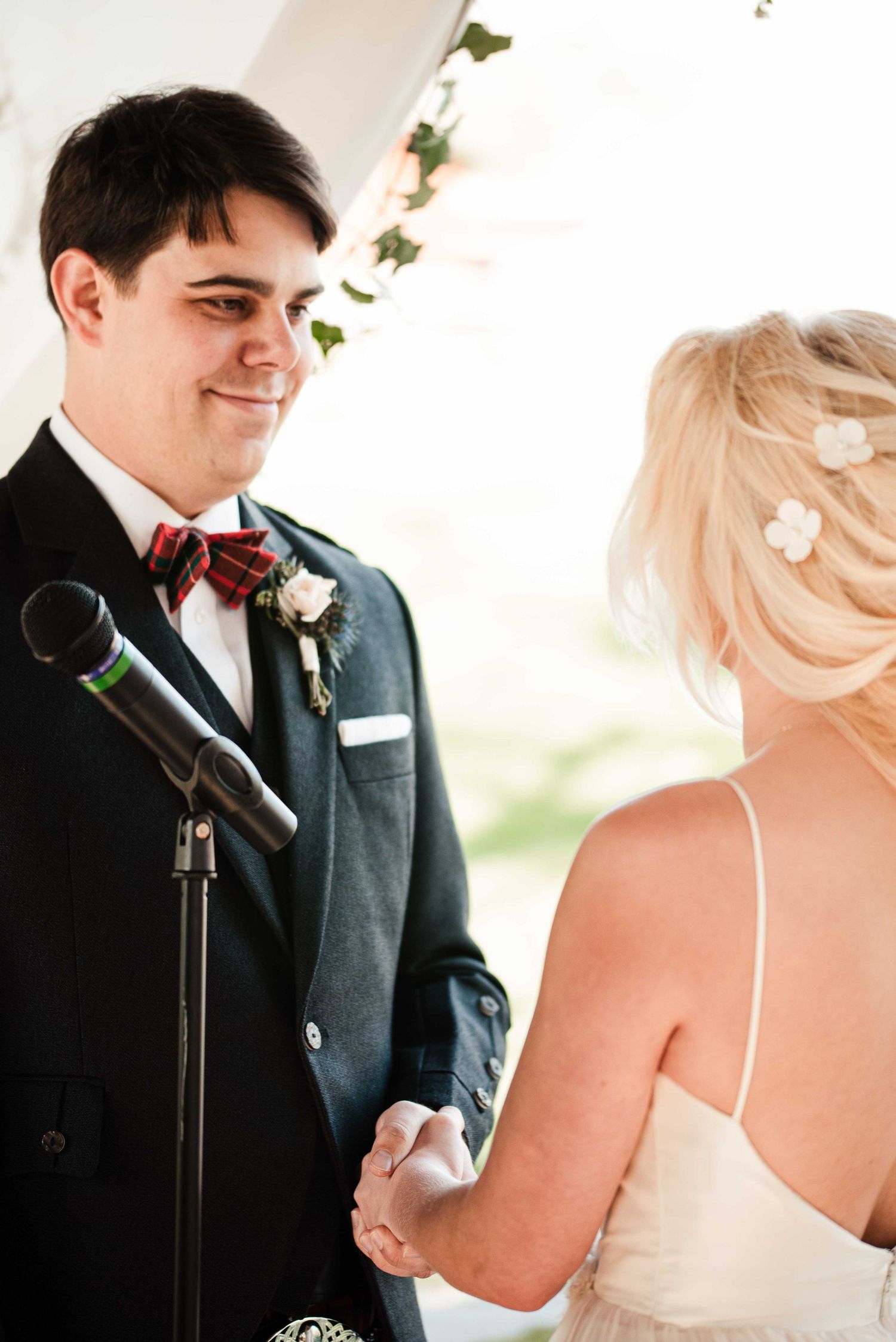 Groom in dark suit and plaid bow tie smiles at blonde bride during outdoor wedding ceremony.