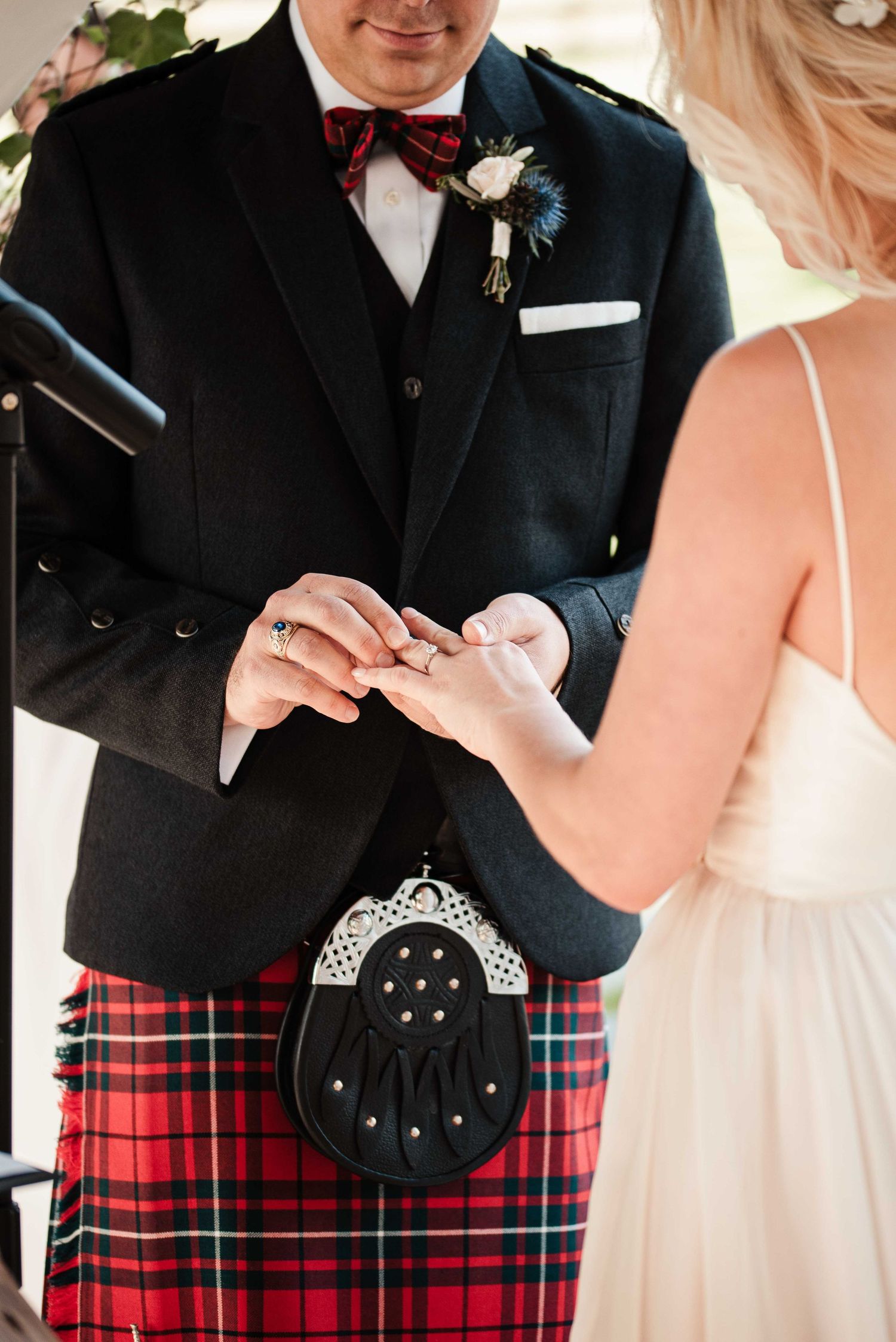 Groom in red tartan kilt and black jacket places wedding ring on bride's finger during Scottish wedding ceremony.