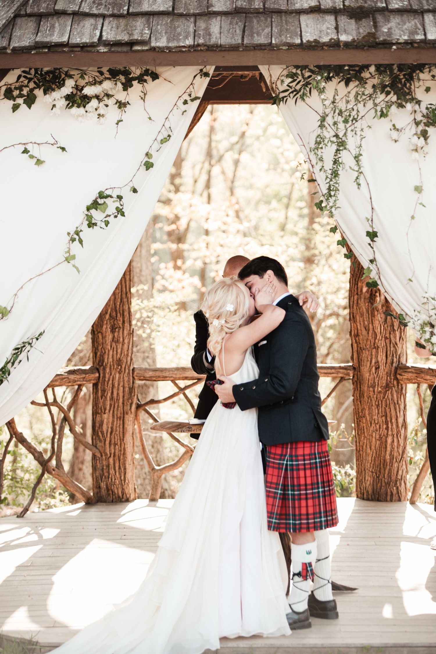 Bride and groom share first kiss under rustic wooden arch with white draping, groom wearing red tartan kilt at outdoor wedding.