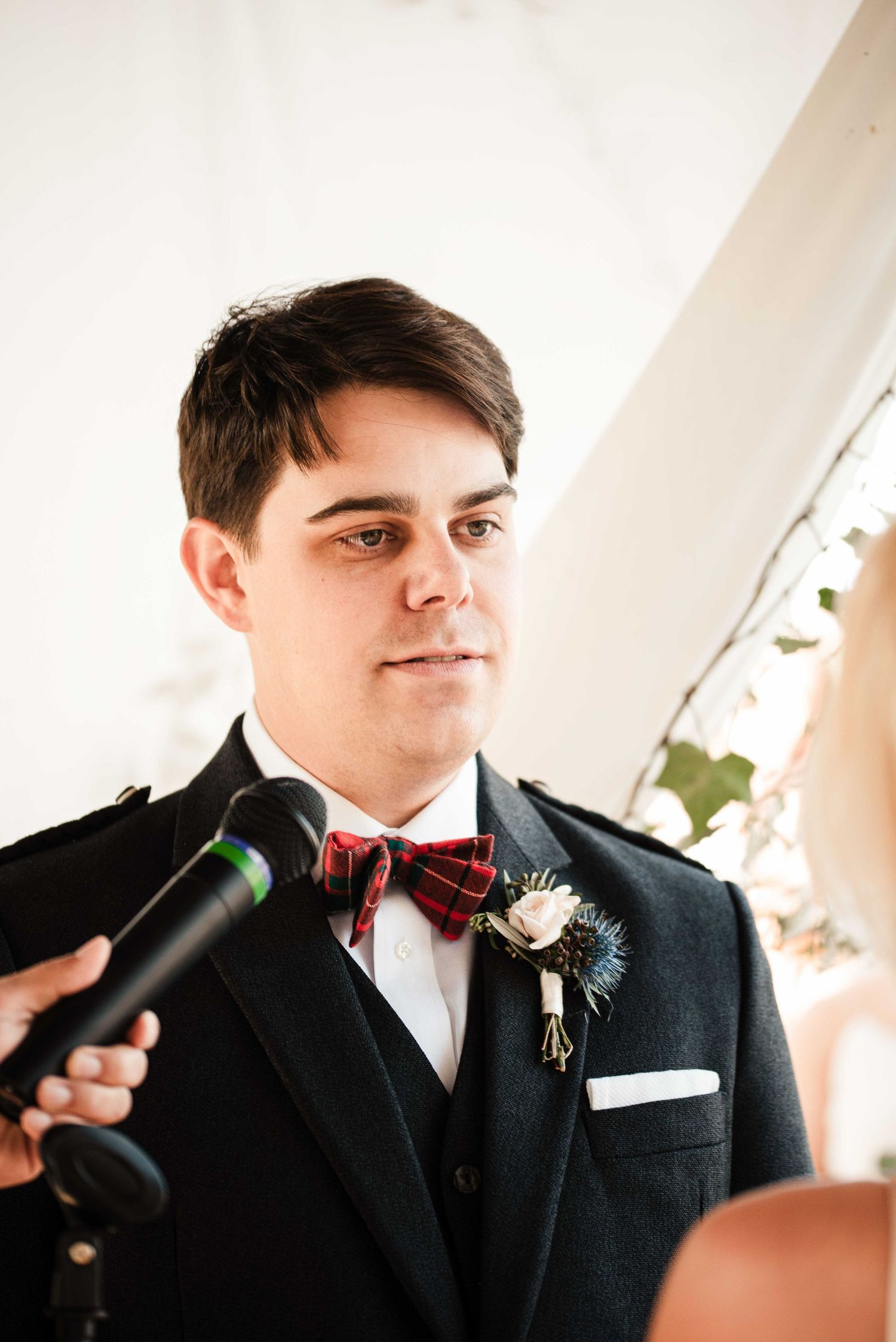 Young groom in tartan bow tie and boutonniere listens attentively during wedding ceremony with microphone nearby.