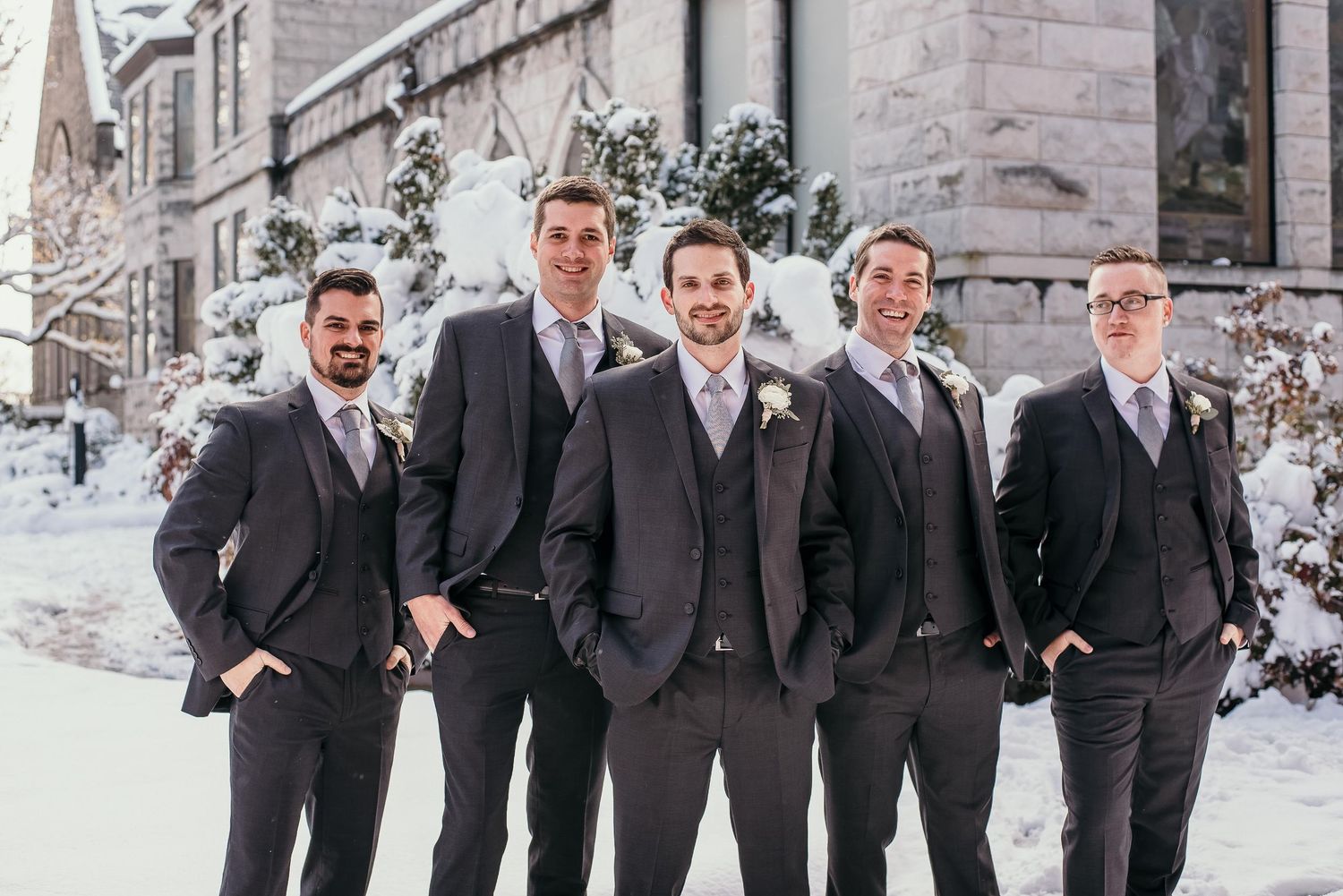 Five groomsmen in dark suits pose outdoors in winter snow near a stone building for a wedding photo.