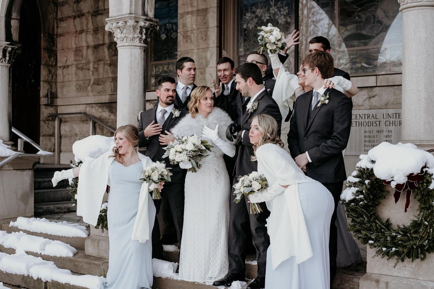 Winter wedding party celebrates on snowy church steps, bridesmaids in white gowns holding bouquets, groomsmen in gray suits cheering.