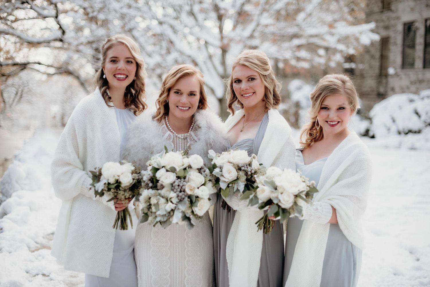 Four bridesmaids in white and gray gowns with fur wraps holding white floral bouquets posing outdoors in a snowy winter setting.
