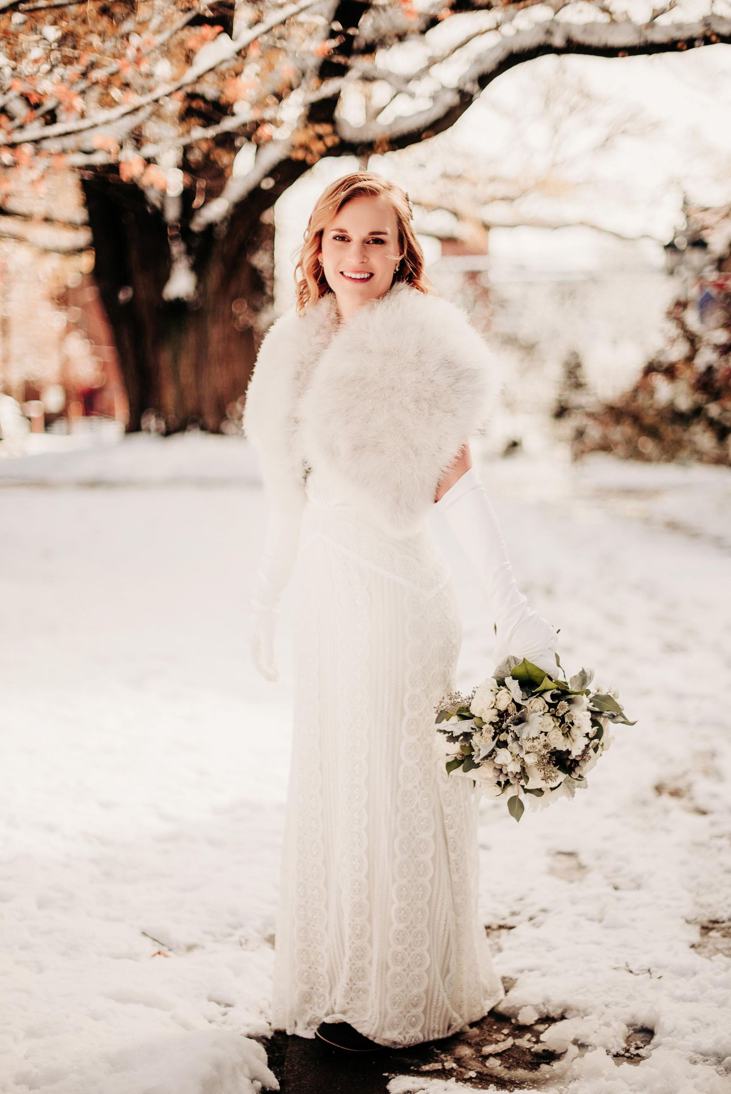 Bride in white lace gown and fur stole holding bouquet, posing in snowy winter outdoor setting.