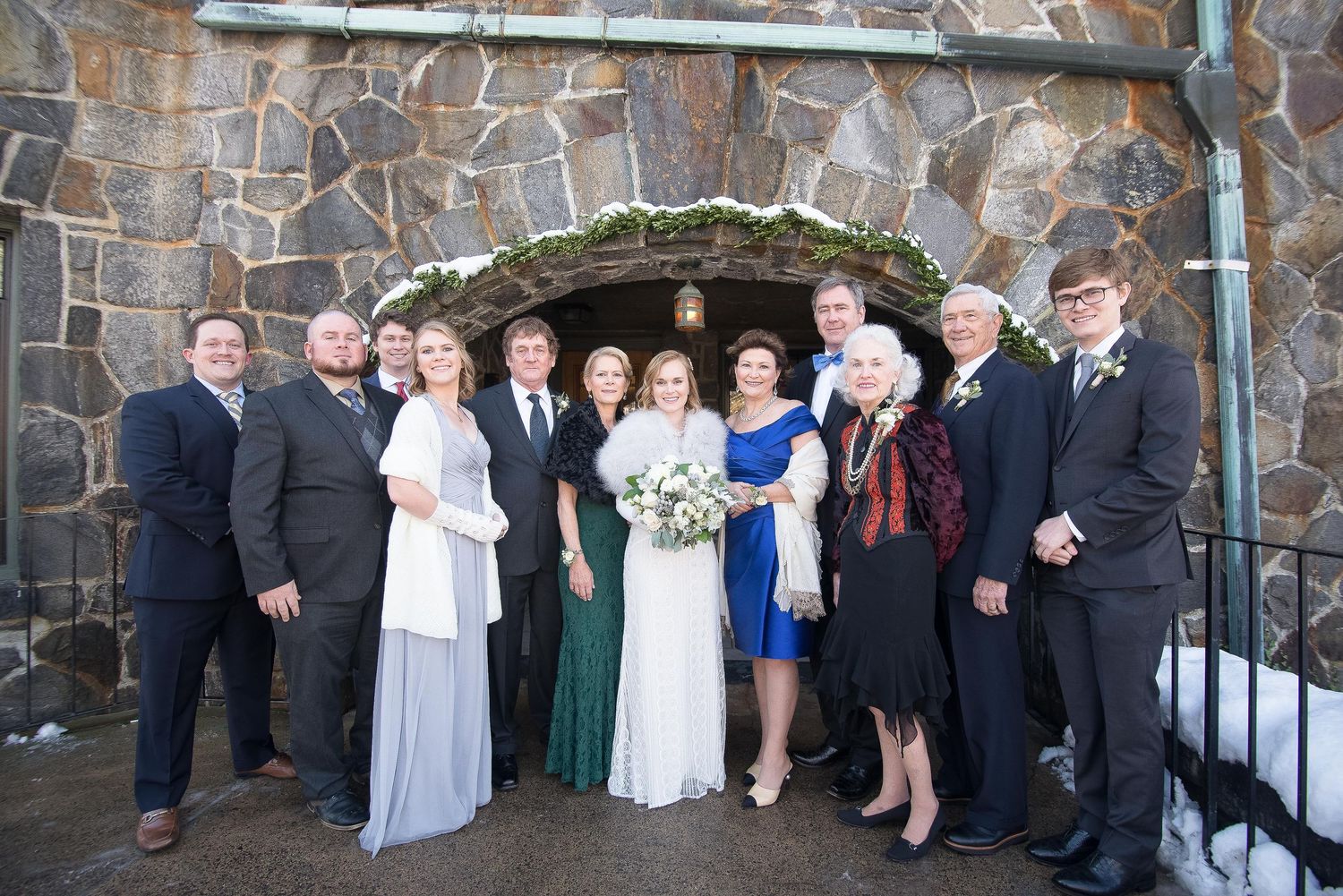 Wedding party posing in formal attire before a stone arch backdrop in winter setting.