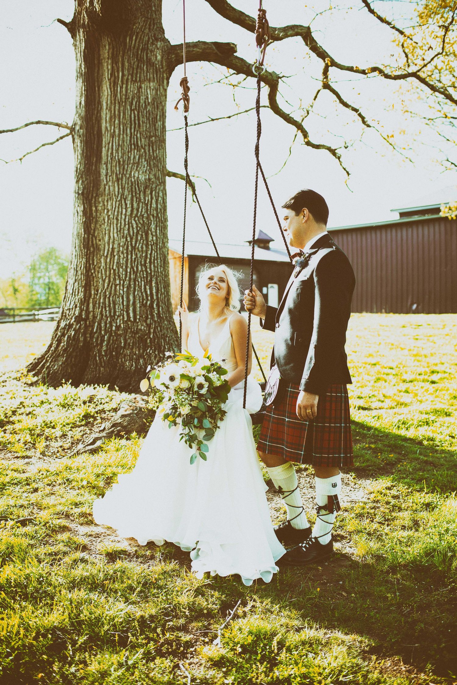 Bride in white gown holding bouquet stands with groom in tartan kilt by a tree swing on a sunny wedding day.