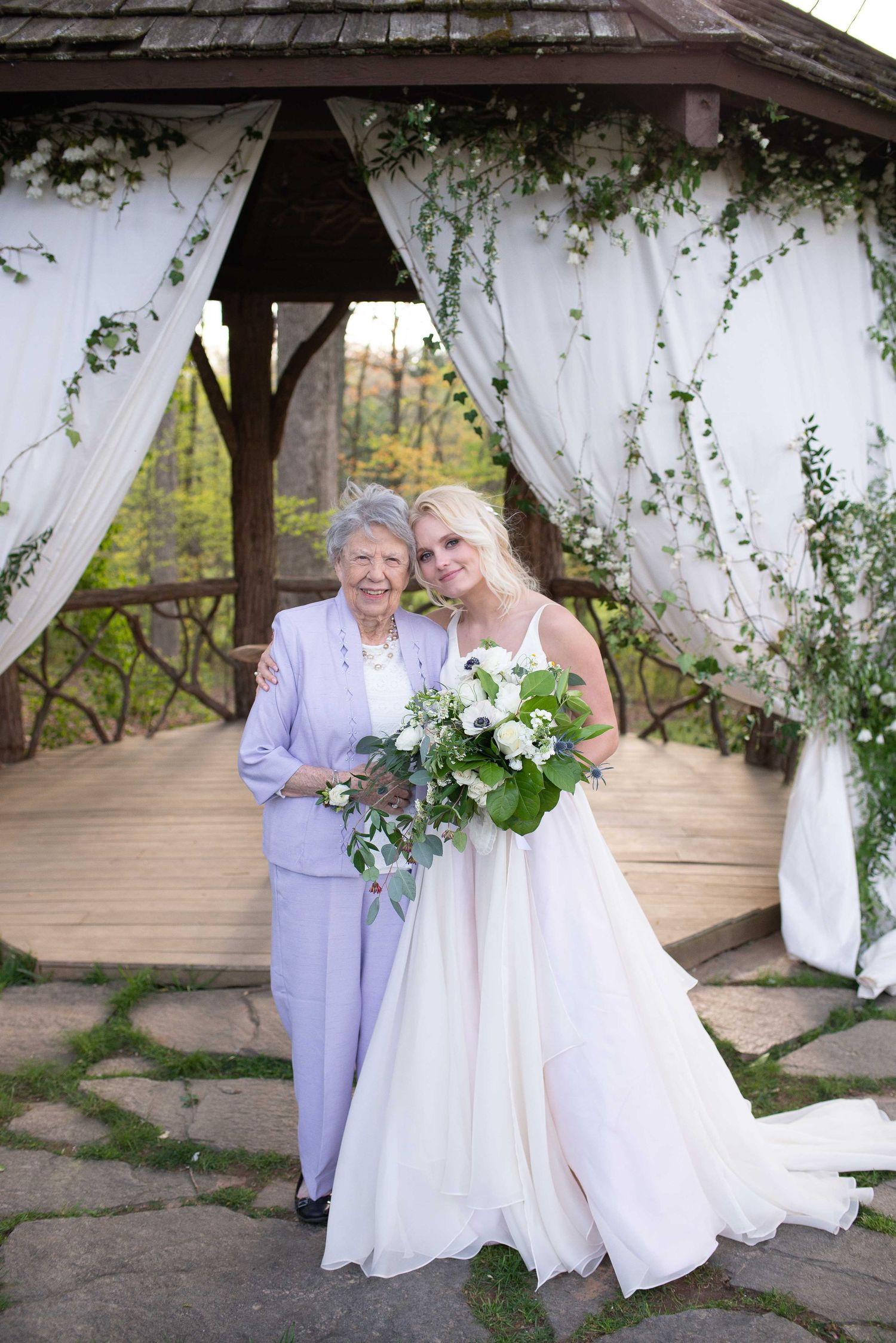 Bride in white gown holding bouquet poses with elderly woman in lavender suit under decorated gazebo.
