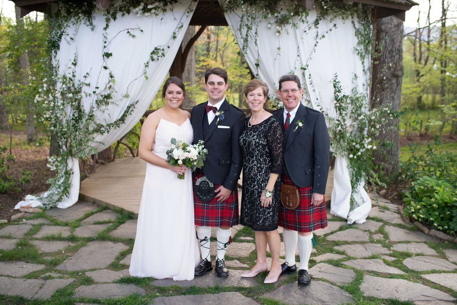 Wedding party poses under floral arbor, bride in white gown, groom and man in traditional Scottish kilts.