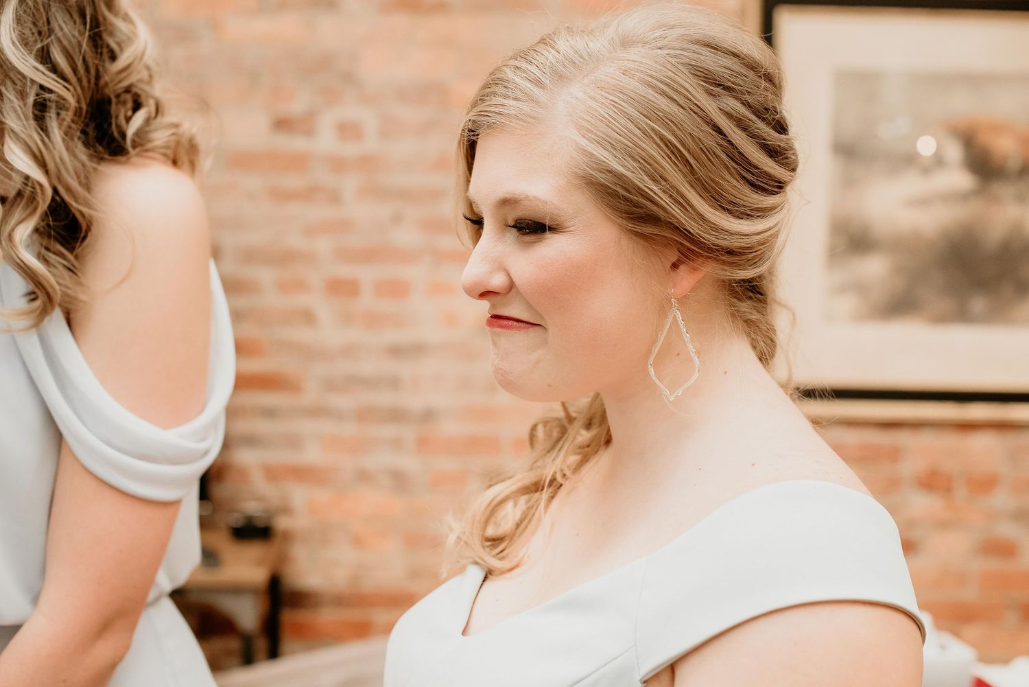 Bride smiling in white dress with elegant updo and drop earrings, standing in a rustic brick-walled venue.