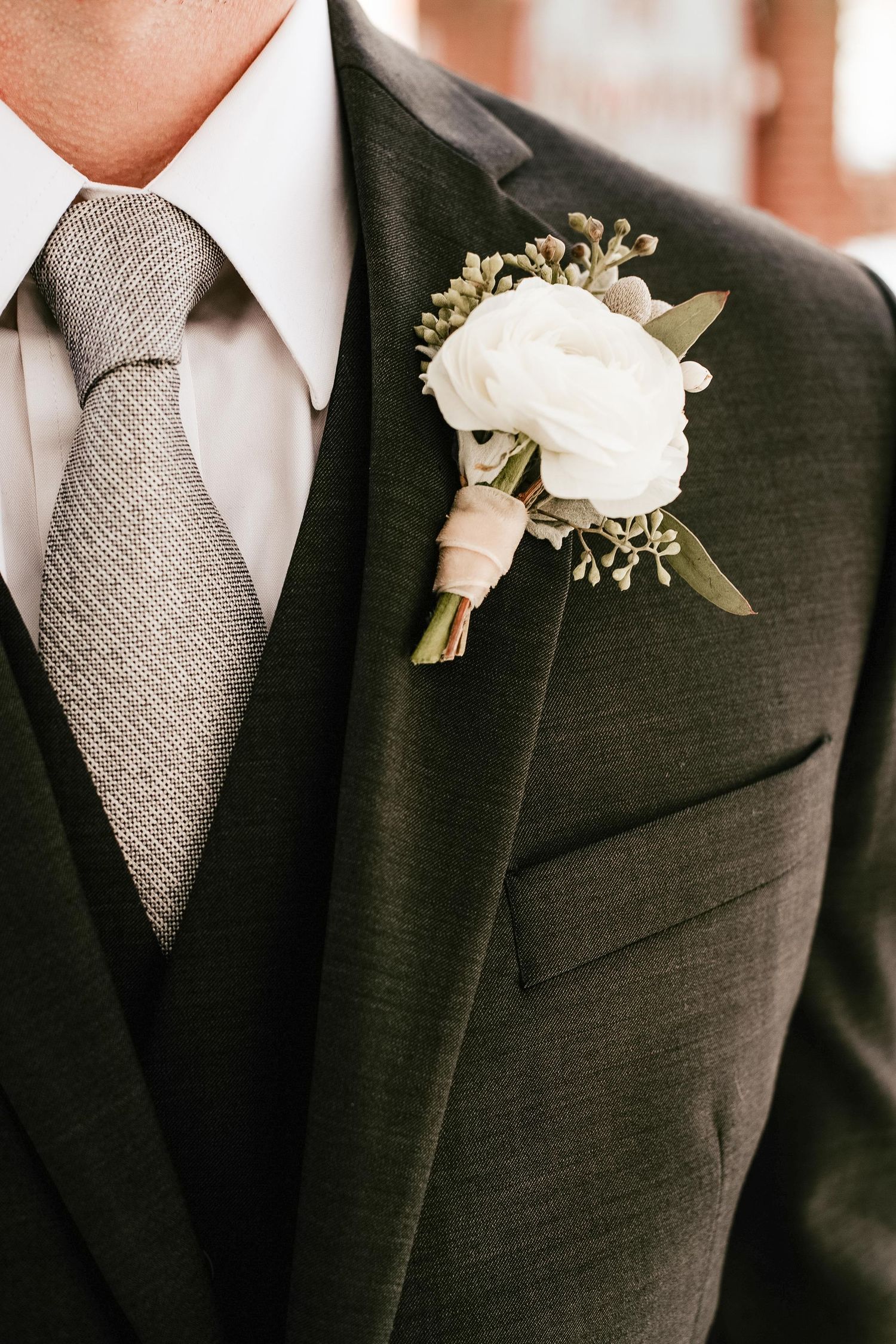 Groom in dark olive three-piece suit with gray tie and white ranunculus boutonniere with greenery.