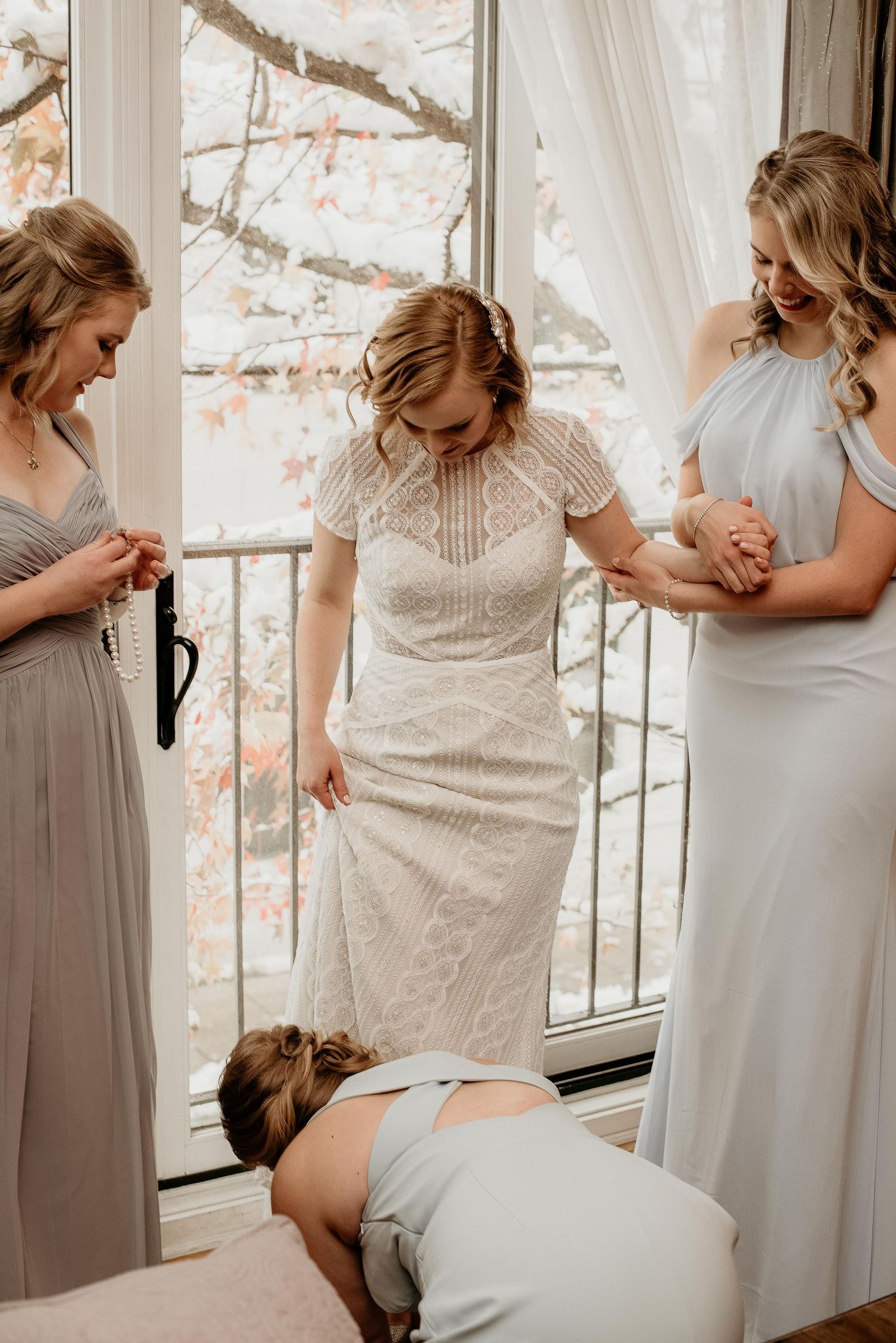 Bridesmaids helping bride into lace wedding dress near window with autumn foliage backdrop.