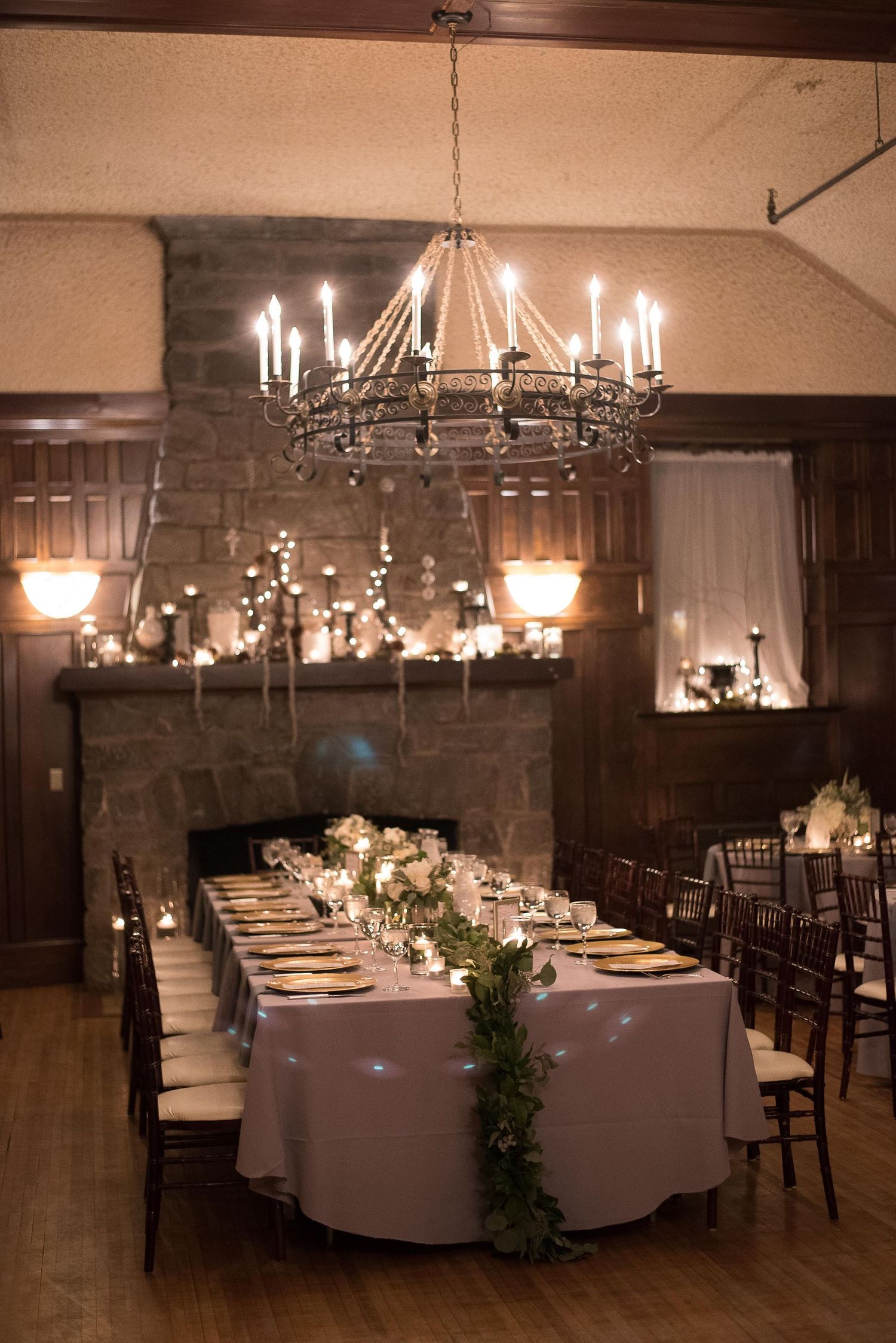 Elegant wedding reception with candlelit chandelier, greenery table runner, and stone fireplace in a warm, intimate venue.