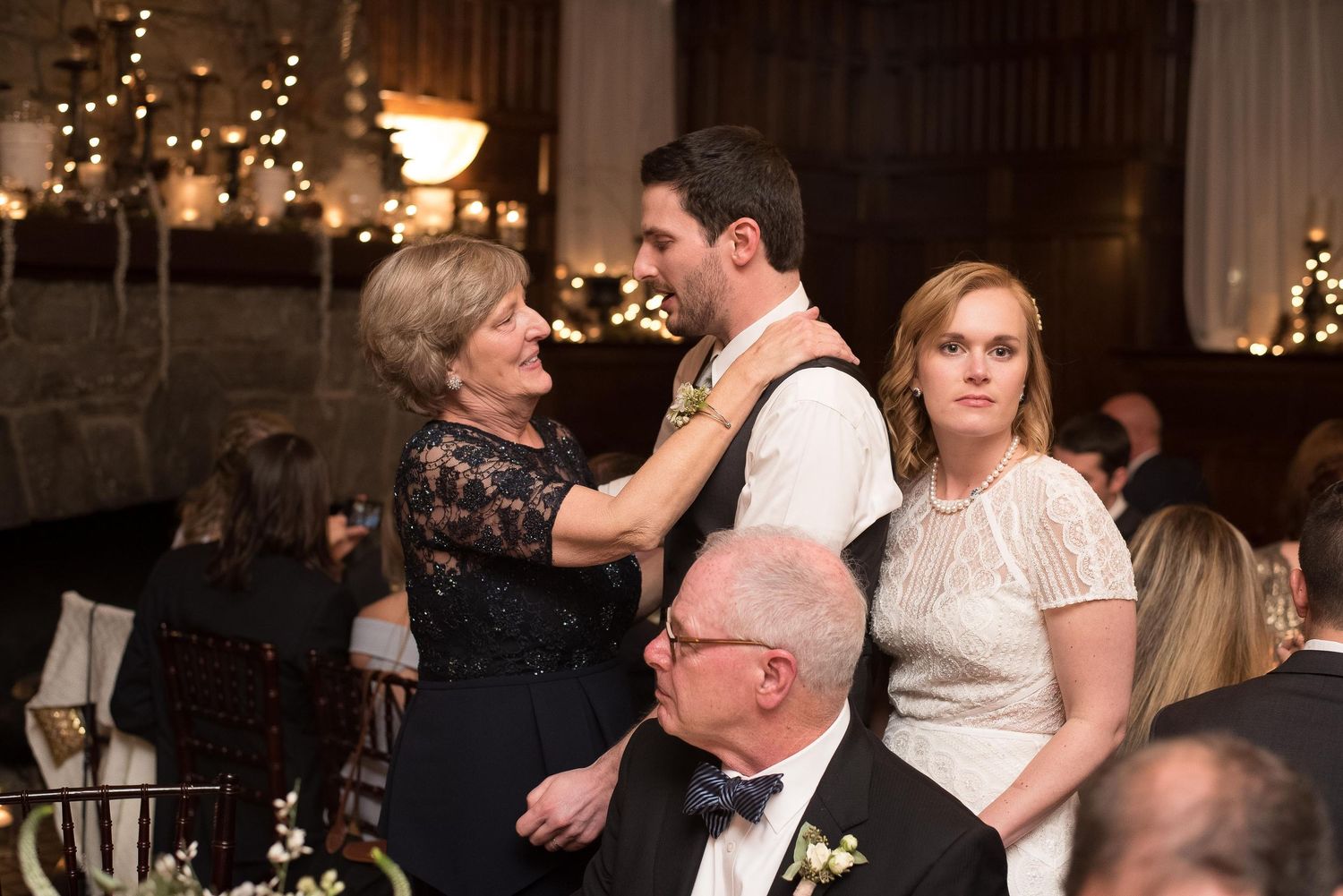 Elderly woman adjusting groom's boutonniere at elegant wedding reception while bride stands nearby.