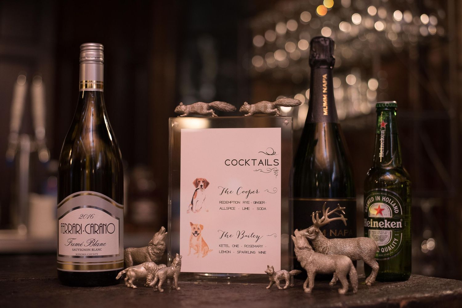 Cocktail menu displayed between wine bottles and Heineken beer, surrounded by small animal figurines on a dark bar surface.