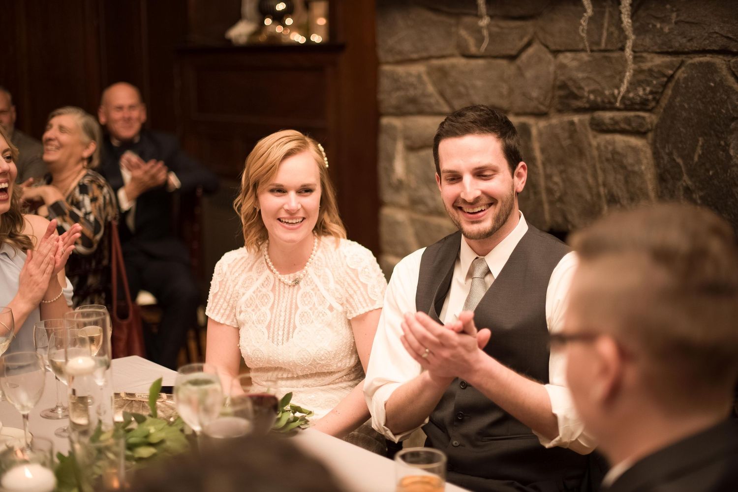 Bride and groom laughing and clapping at wedding reception dinner table in rustic venue.