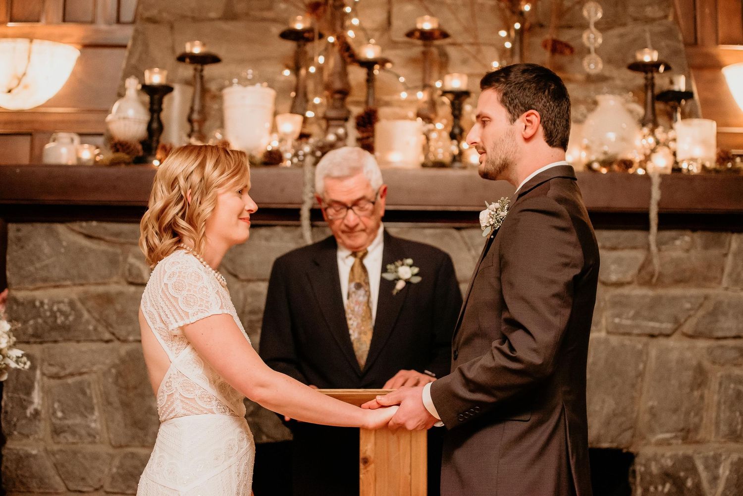 Bride and groom hold hands during intimate wedding ceremony before stone fireplace with candles.