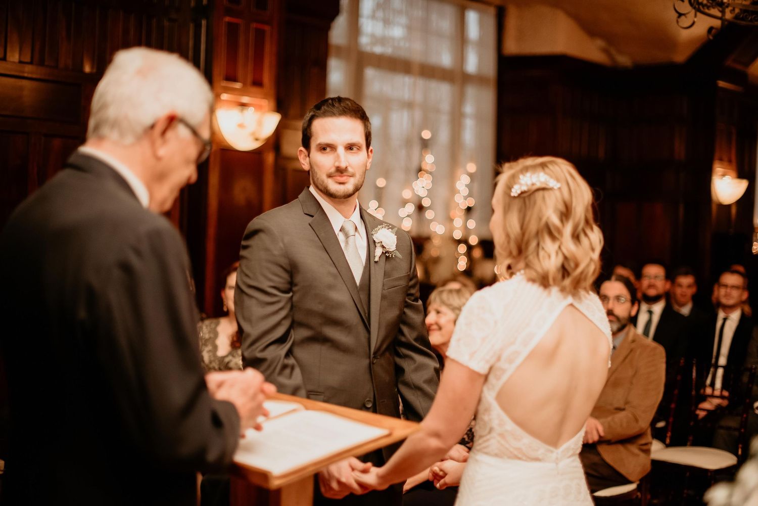 Groom smiling at bride during indoor wedding ceremony, officiant holding book, guests seated in elegant wood-paneled venue.