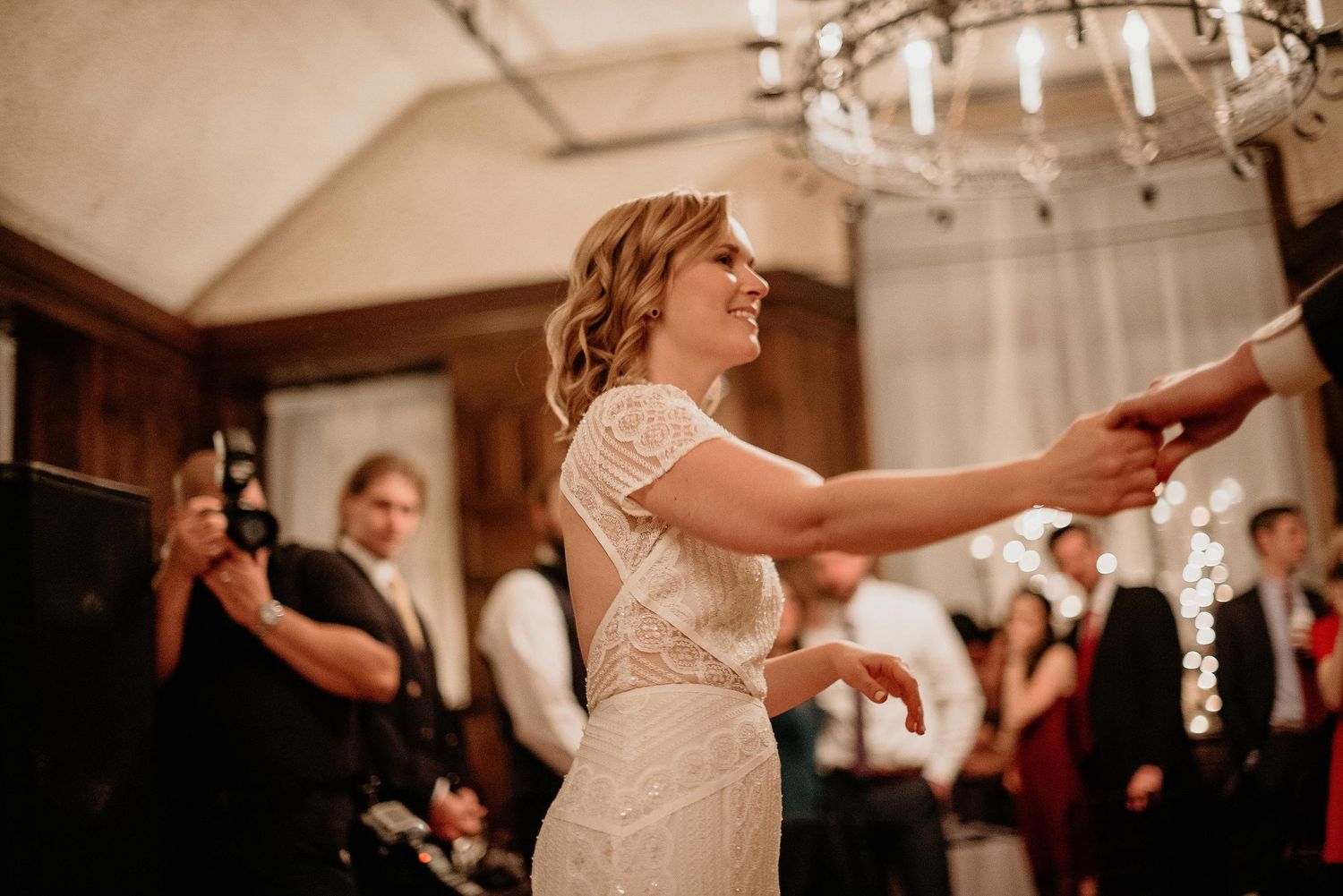 Bride in beaded white gown dancing and smiling joyfully at elegant wedding reception with chandelier.