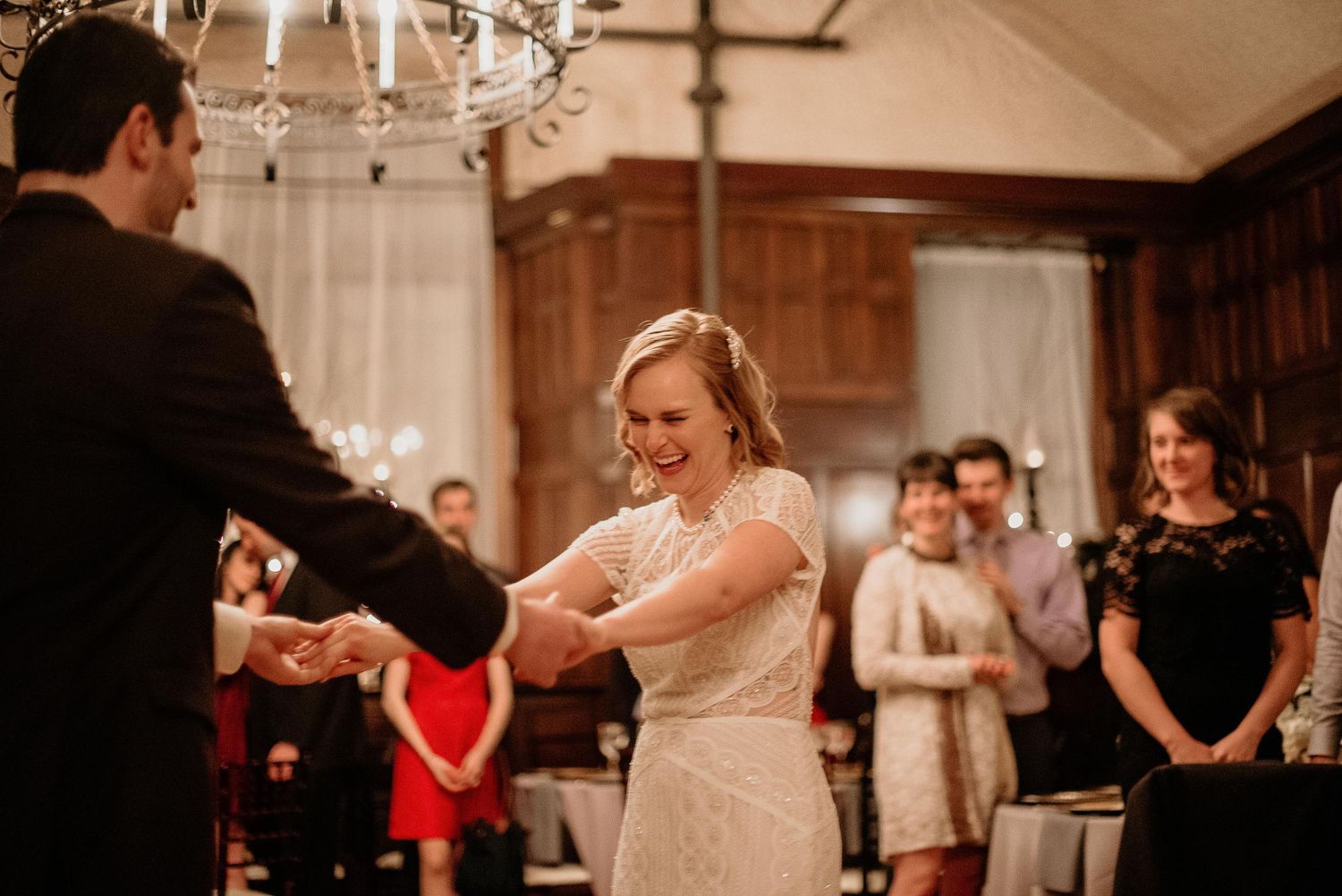 Bride laughing joyfully during first dance with groom at elegant wedding reception with chandelier.