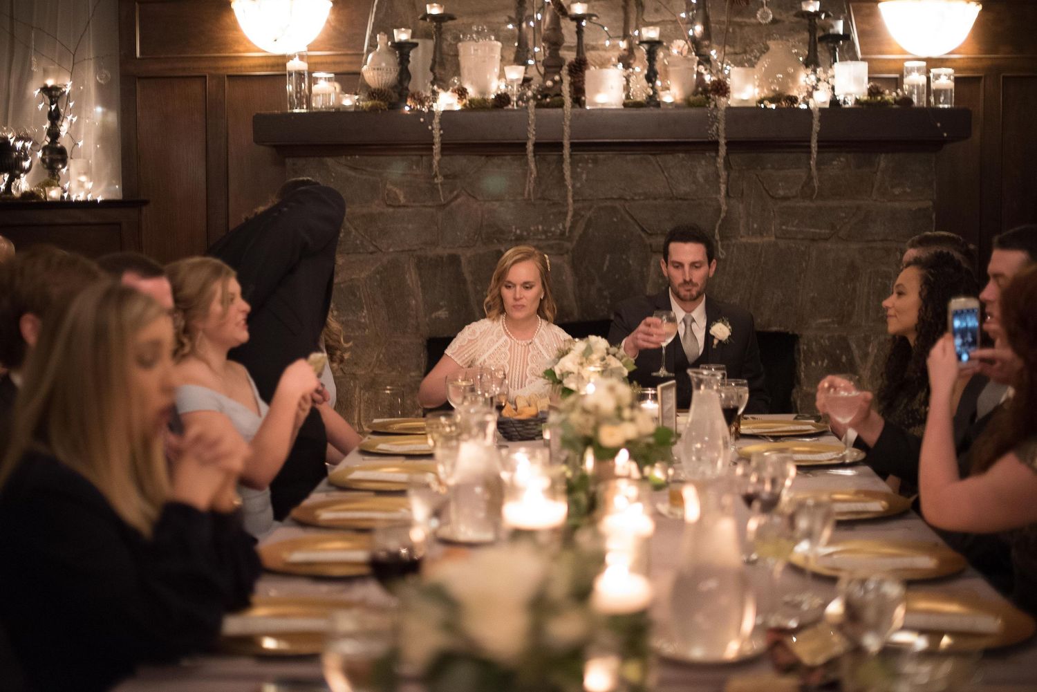 Bride and groom seated at elegant candlelit wedding reception dinner table surrounded by guests in a cozy rustic venue.