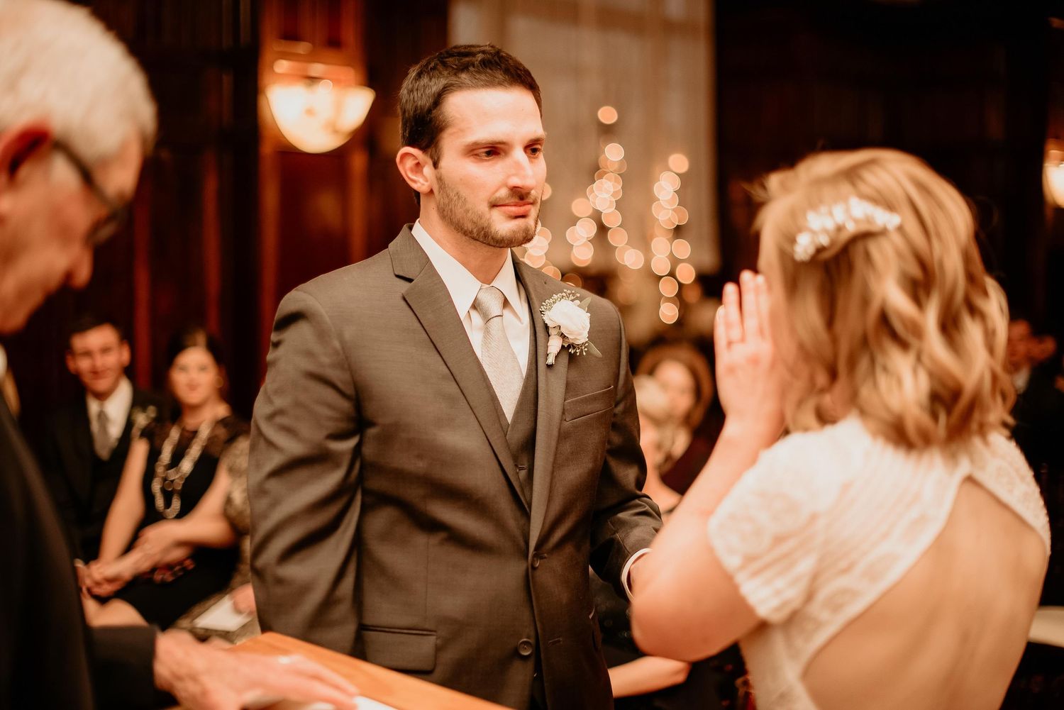 Groom in gray suit watches bride recite vows during elegant indoor wedding ceremony with warm bokeh lighting.