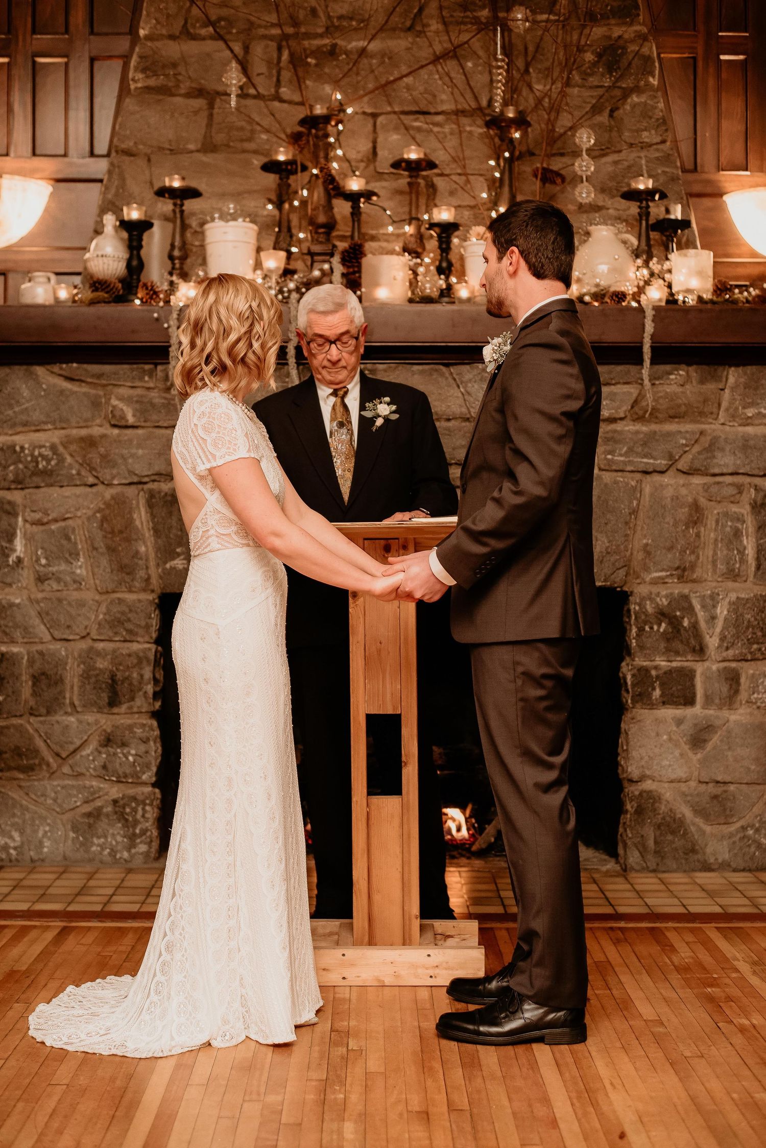 Bride and groom exchange vows at rustic stone fireplace altar with candles and an officiant during indoor wedding ceremony.