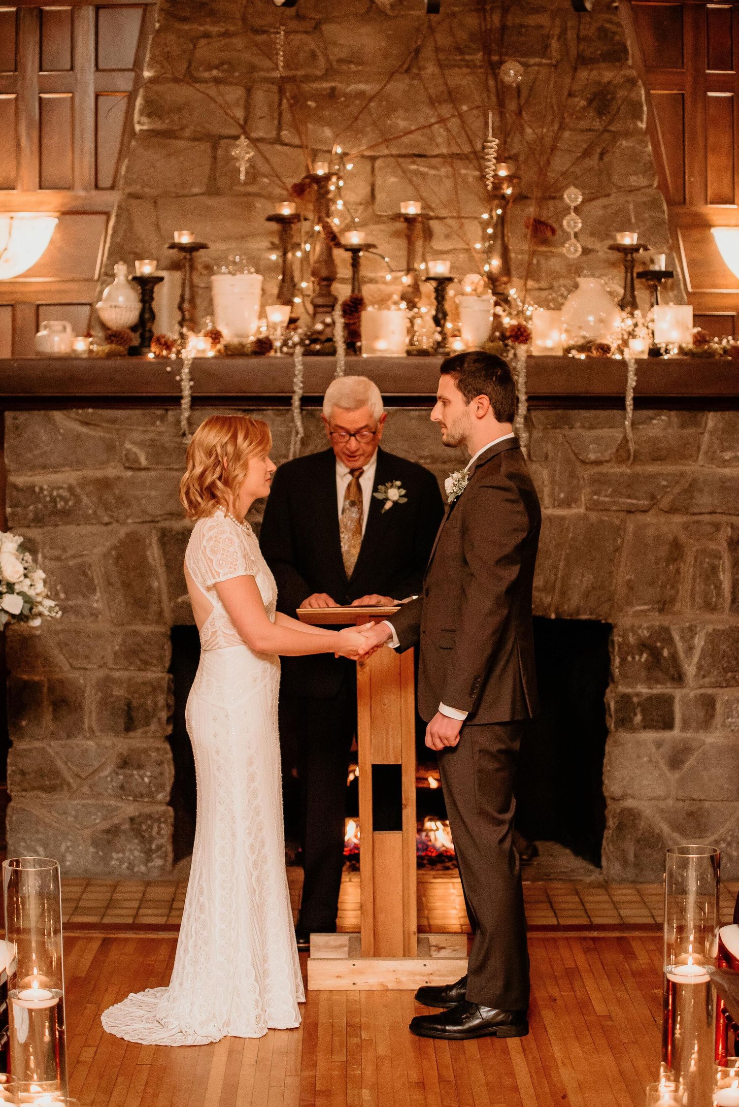 Bride and groom exchange vows at candlelit indoor wedding ceremony before a stone fireplace with an officiant.