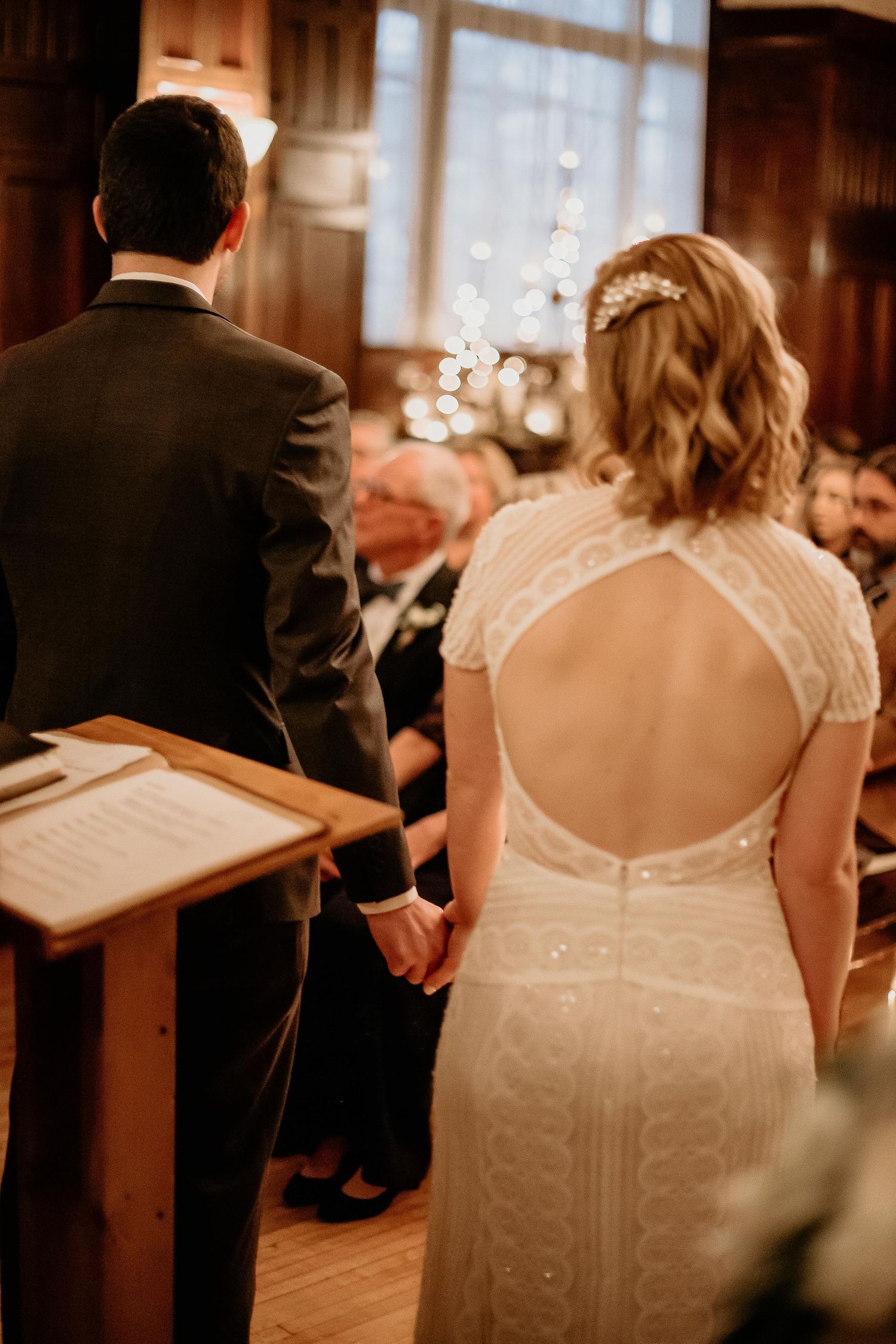 Bride and groom holding hands at wedding altar, bride in elegant open-back lace dress with hair accessory.