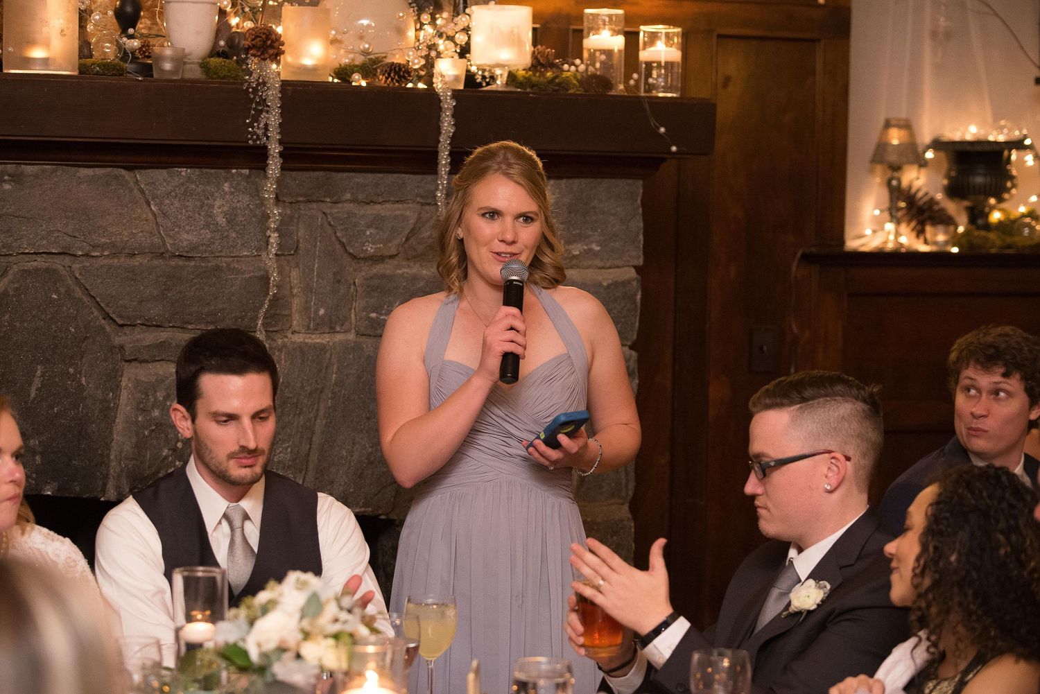 Bridesmaid in gray dress giving a speech at a wedding reception near a stone fireplace.