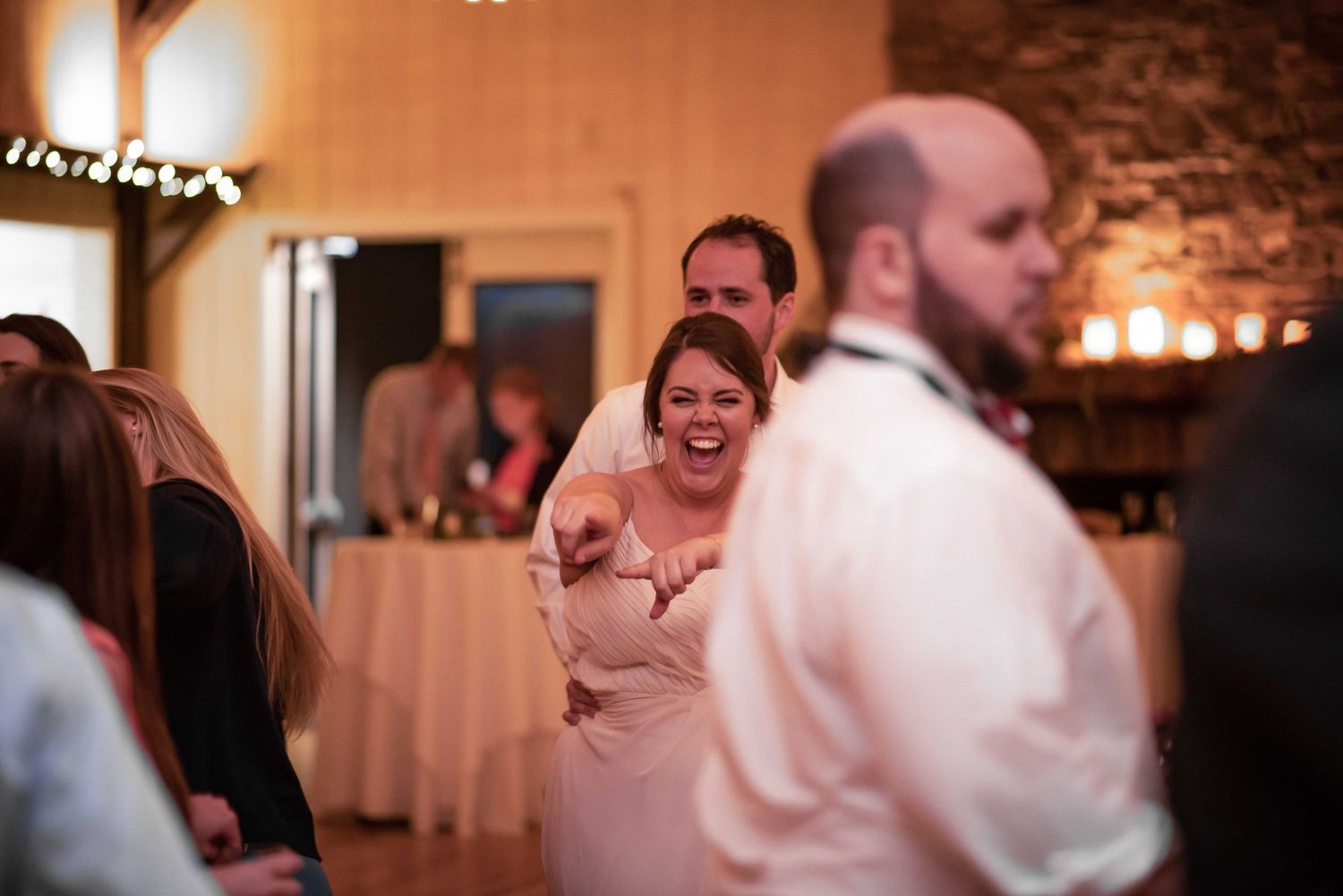 Bride laughing and pointing joyfully with groom behind her during lively wedding reception in rustic venue.
