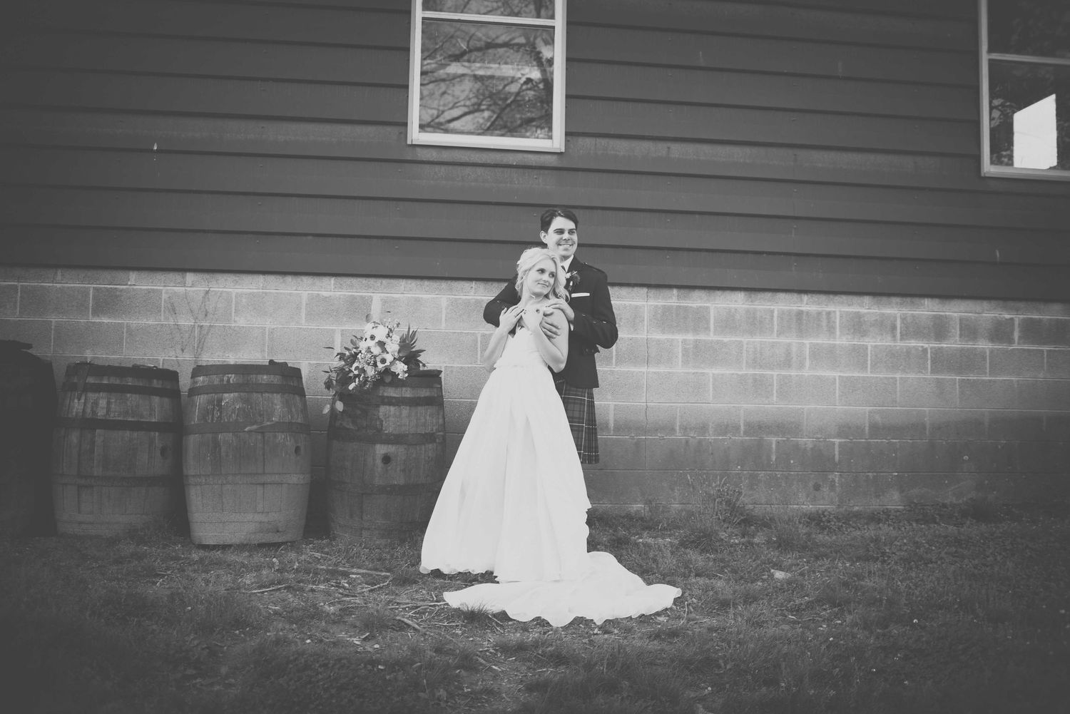 Black and white photo of a bride and groom posing outside a rustic barn venue with wine barrels on their wedding day.