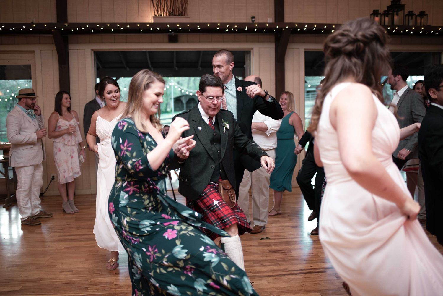 Guests dancing energetically at a wedding reception, including a man in a tartan kilt in a barn venue.