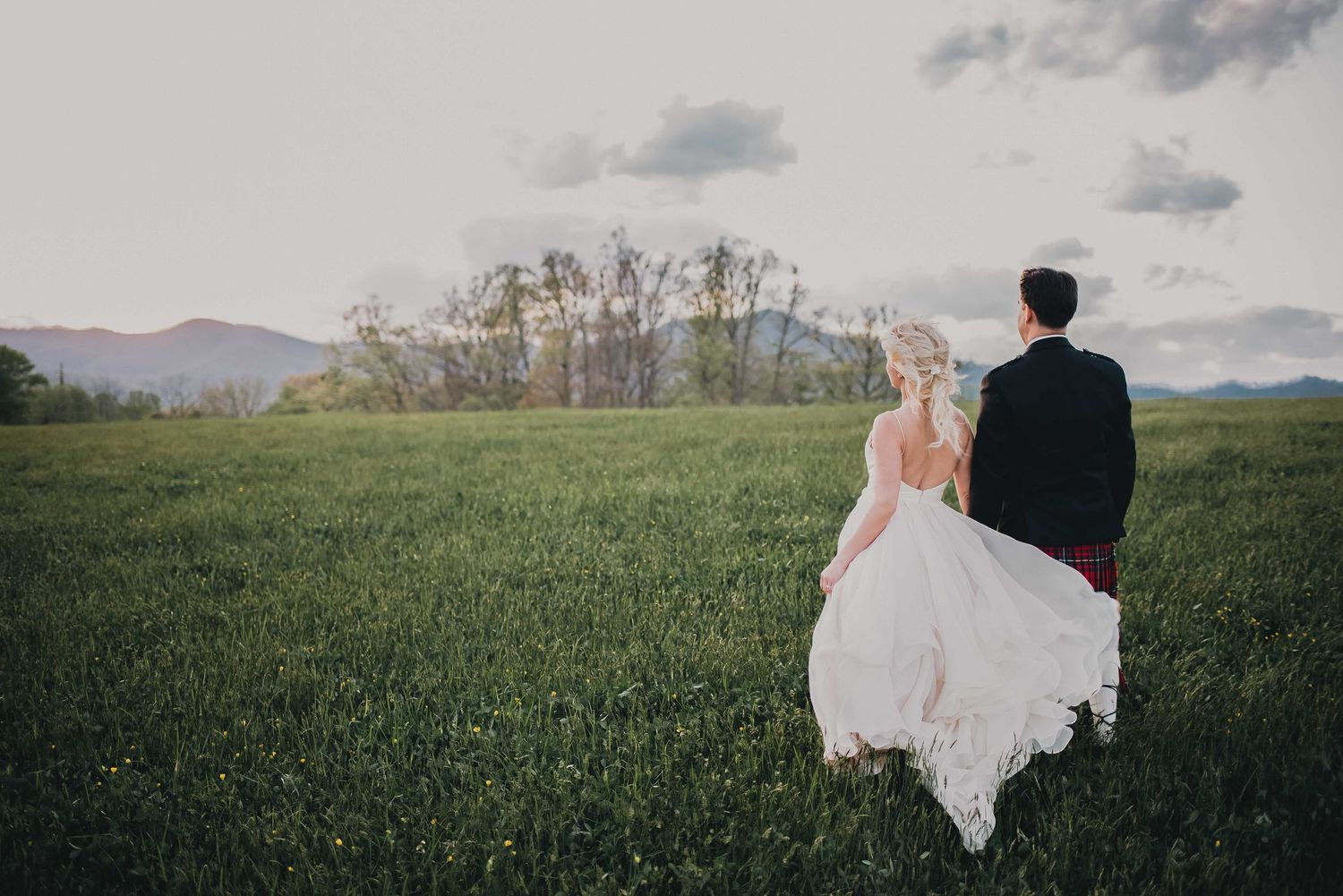 Bride and groom walking through lush green field with mountain backdrop at sunset wedding.