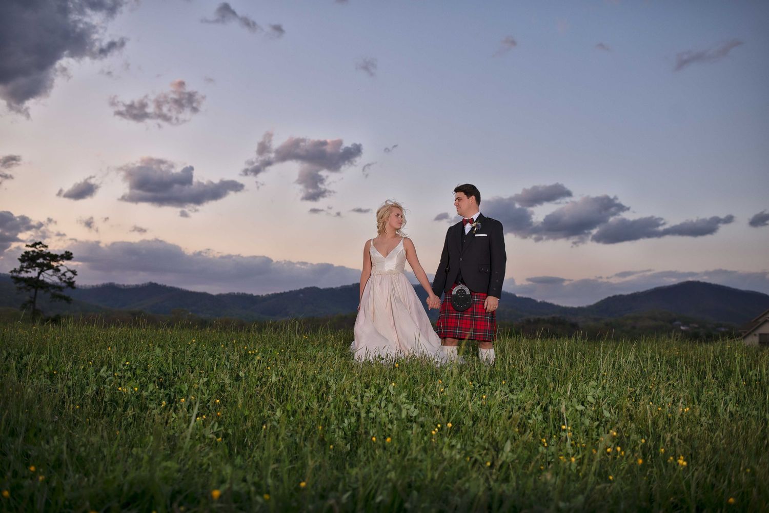 Bride and groom in kilt standing in green field with mountain sunset backdrop.