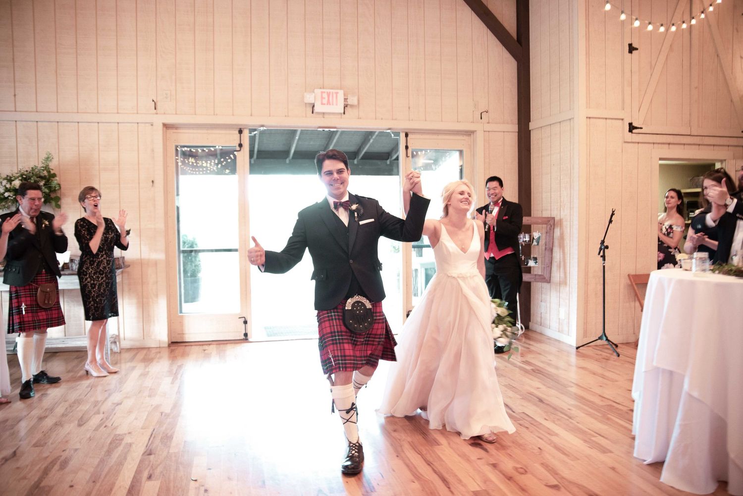 Groom in tartan kilt and bride in white gown make grand entrance at wedding reception as guests applaud.