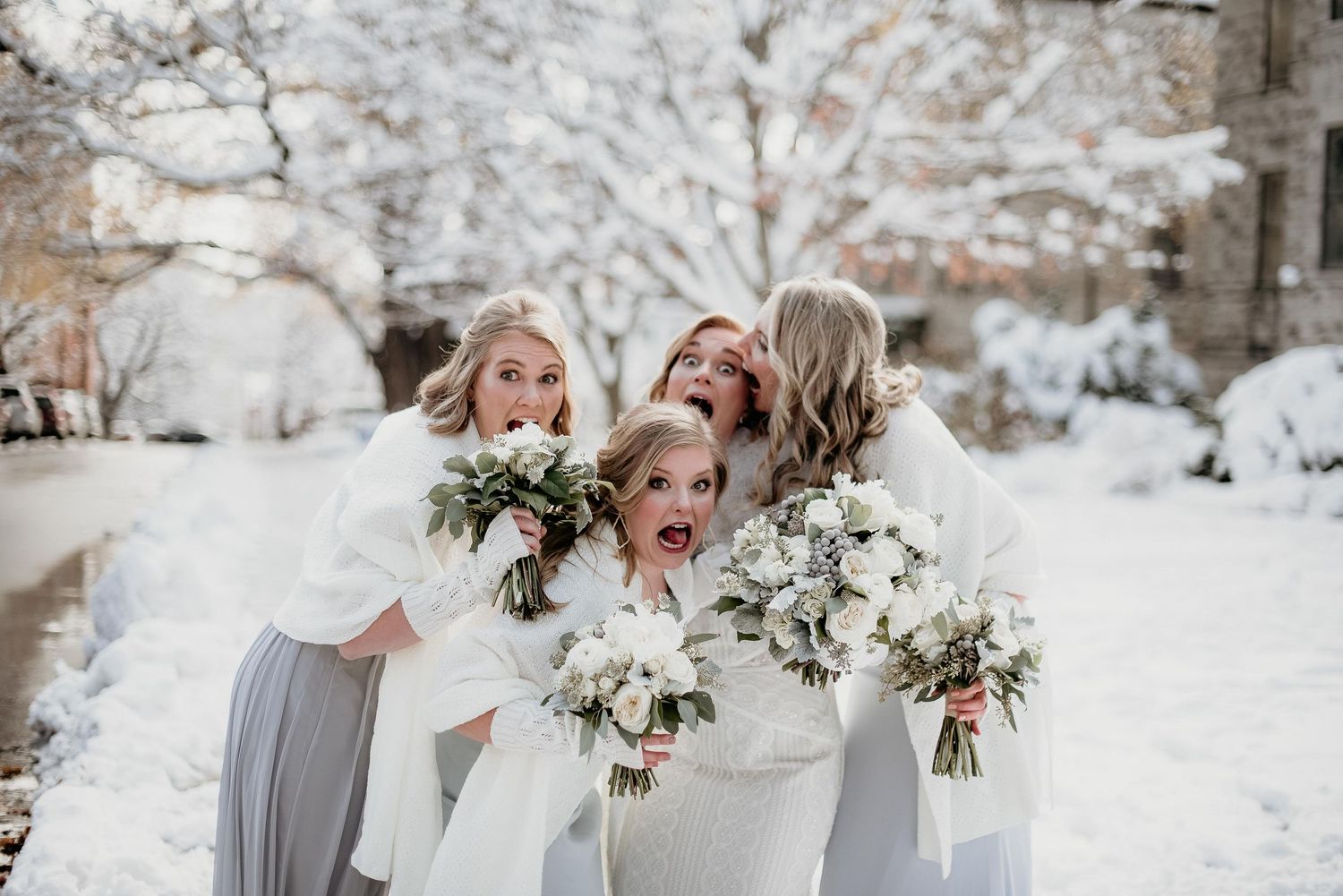 Four bridesmaids in white wraps holding floral bouquets posing playfully in a snowy winter wedding outdoor setting.