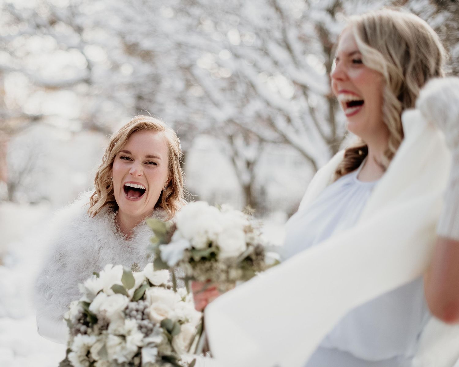 Two brides laughing joyfully outdoors in snow, holding white bouquets during a winter wedding.