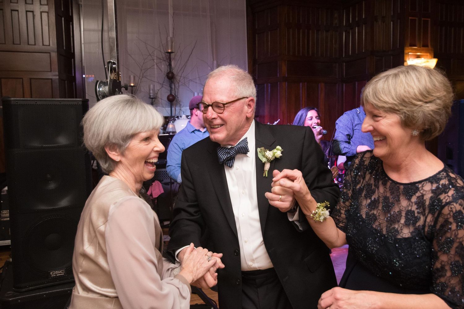 Three elegantly dressed seniors laughing and dancing together at a formal wedding reception event.