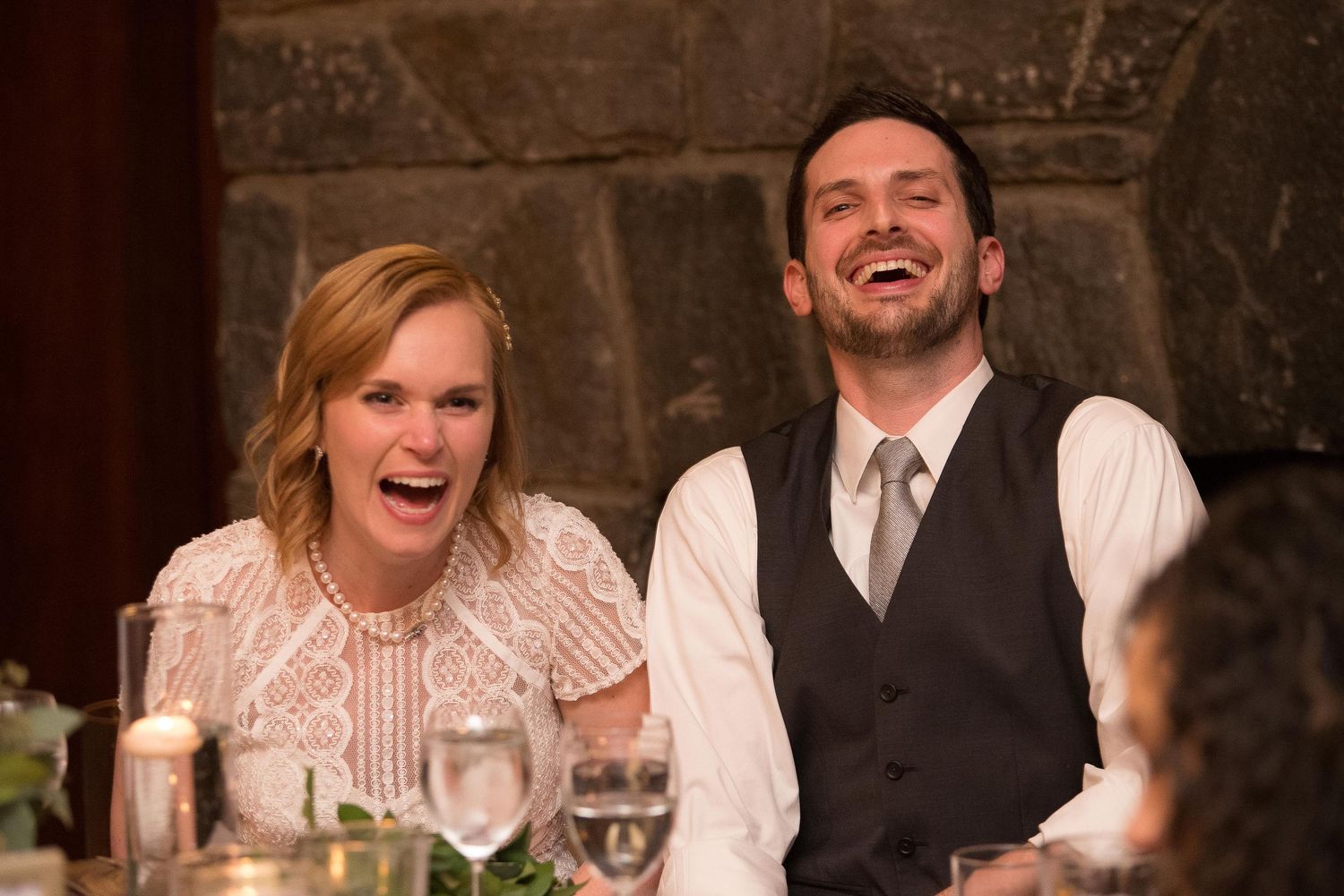 A man and woman laugh joyfully together at a wedding reception dinner, seated at an elegantly set table.