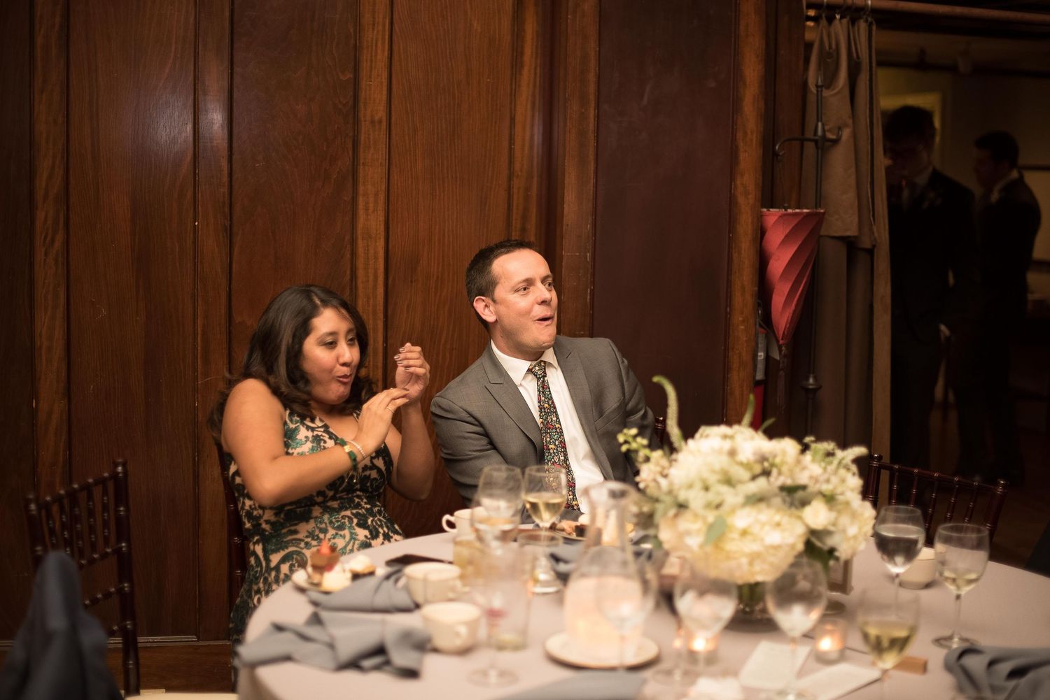 Couple laughing and clapping at an elegant wedding reception dinner table with white floral centerpiece.