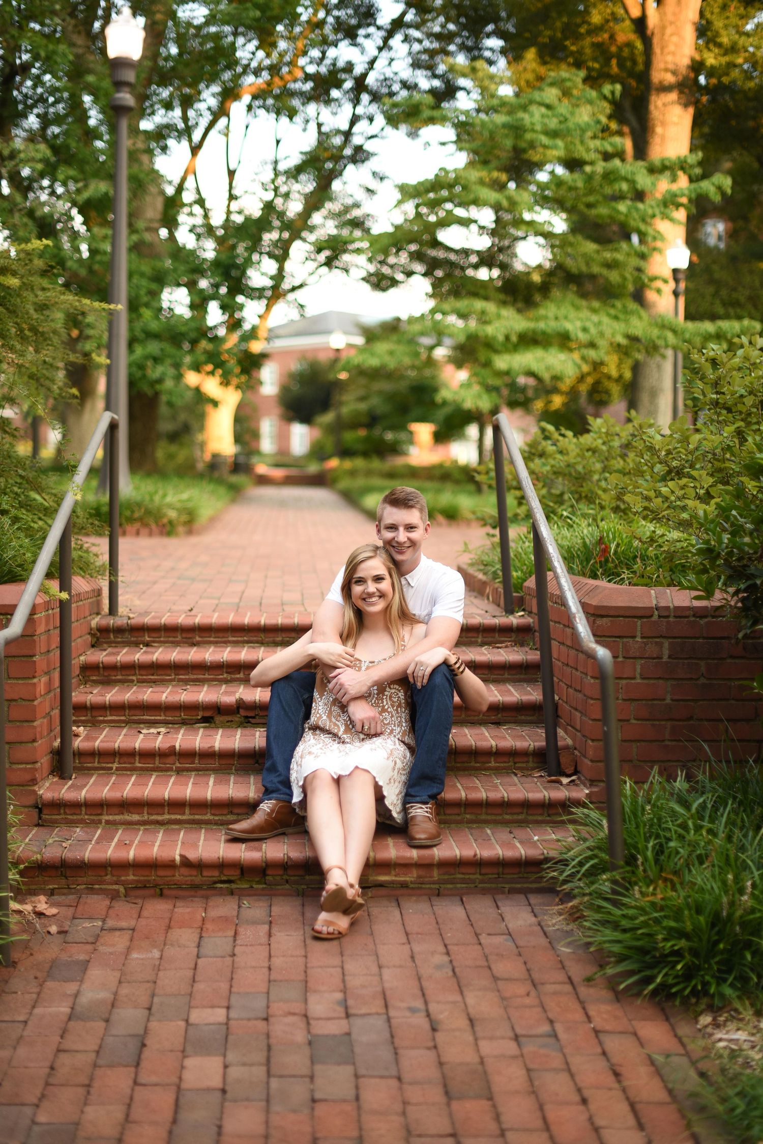 Couple sitting together on brick steps in a lush garden pathway, smiling warmly during an engagement photo session.