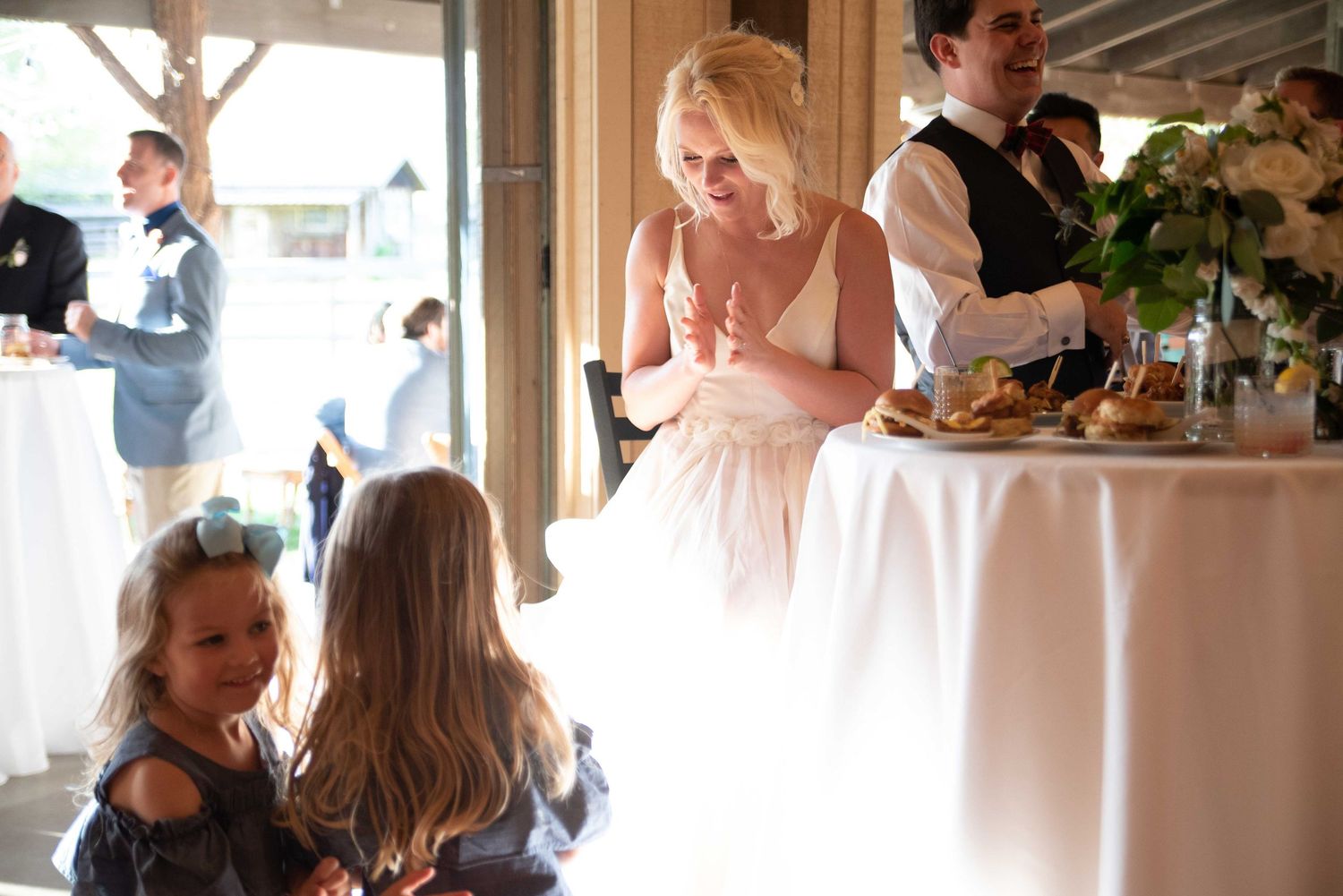 Bride in white gown interacts with young flower girls at elegant wedding reception table.